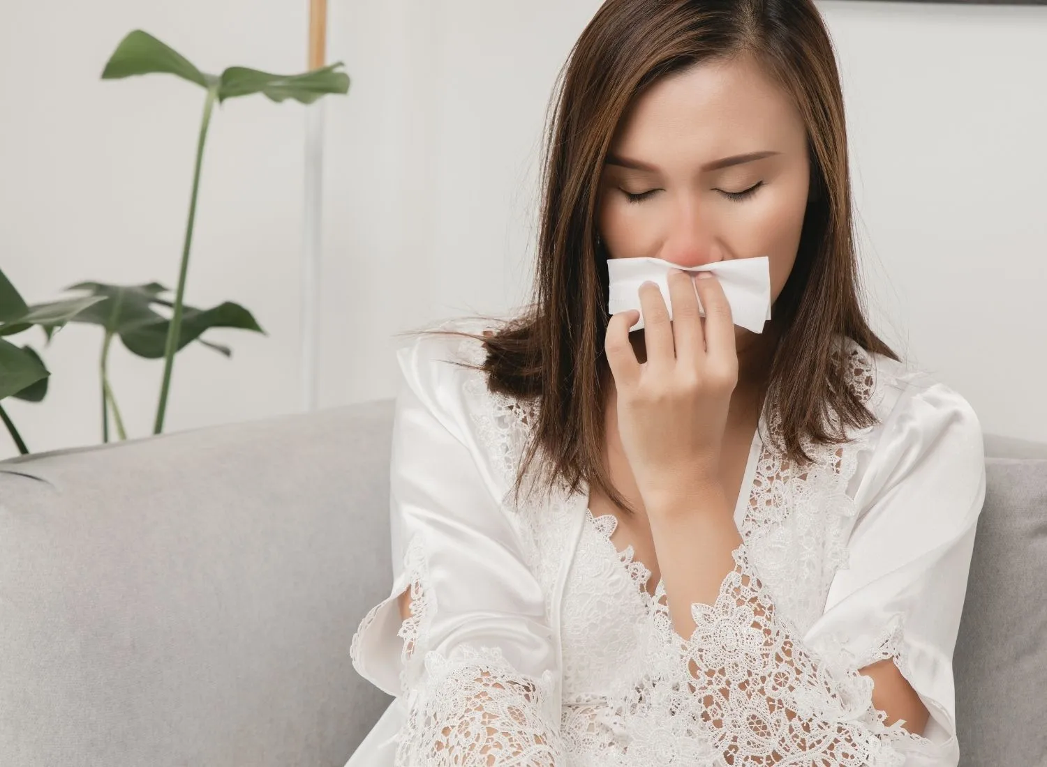 Woman with closed eyes holding tissue to nose, sitting on a sofa with plants in background.