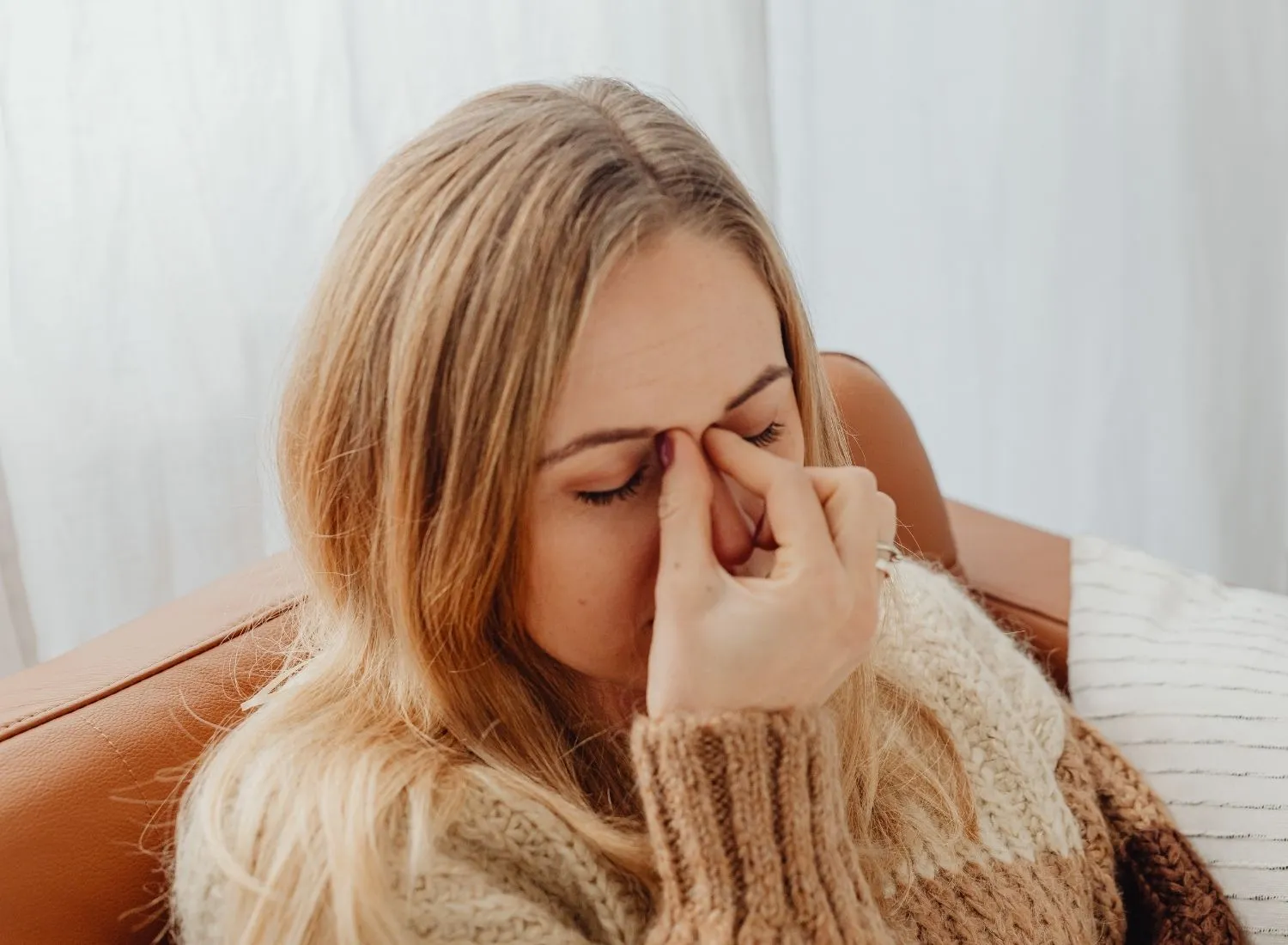 Woman in knit sweater sitting on couch, pinching the bridge of her nose with eyes closed.