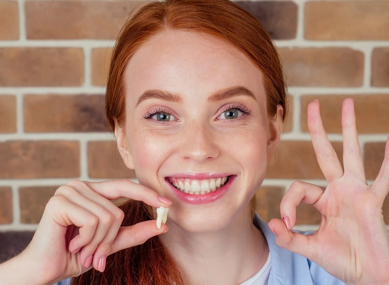 Smiling woman with red hair holding a tooth in one hand and making an okay sign with the other.