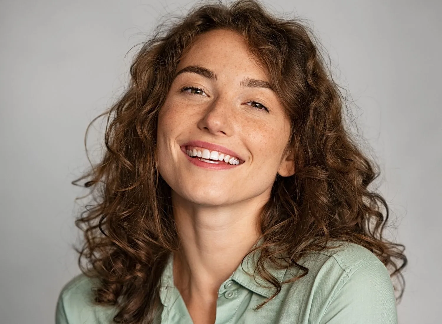 Smiling woman with curly brown hair and freckles wearing a light green shirt against gray background.