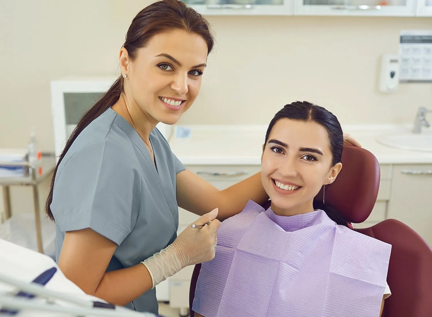 Dentist in scrubs with gloves smiles next to a female patient seated in a dental chair with a bib.