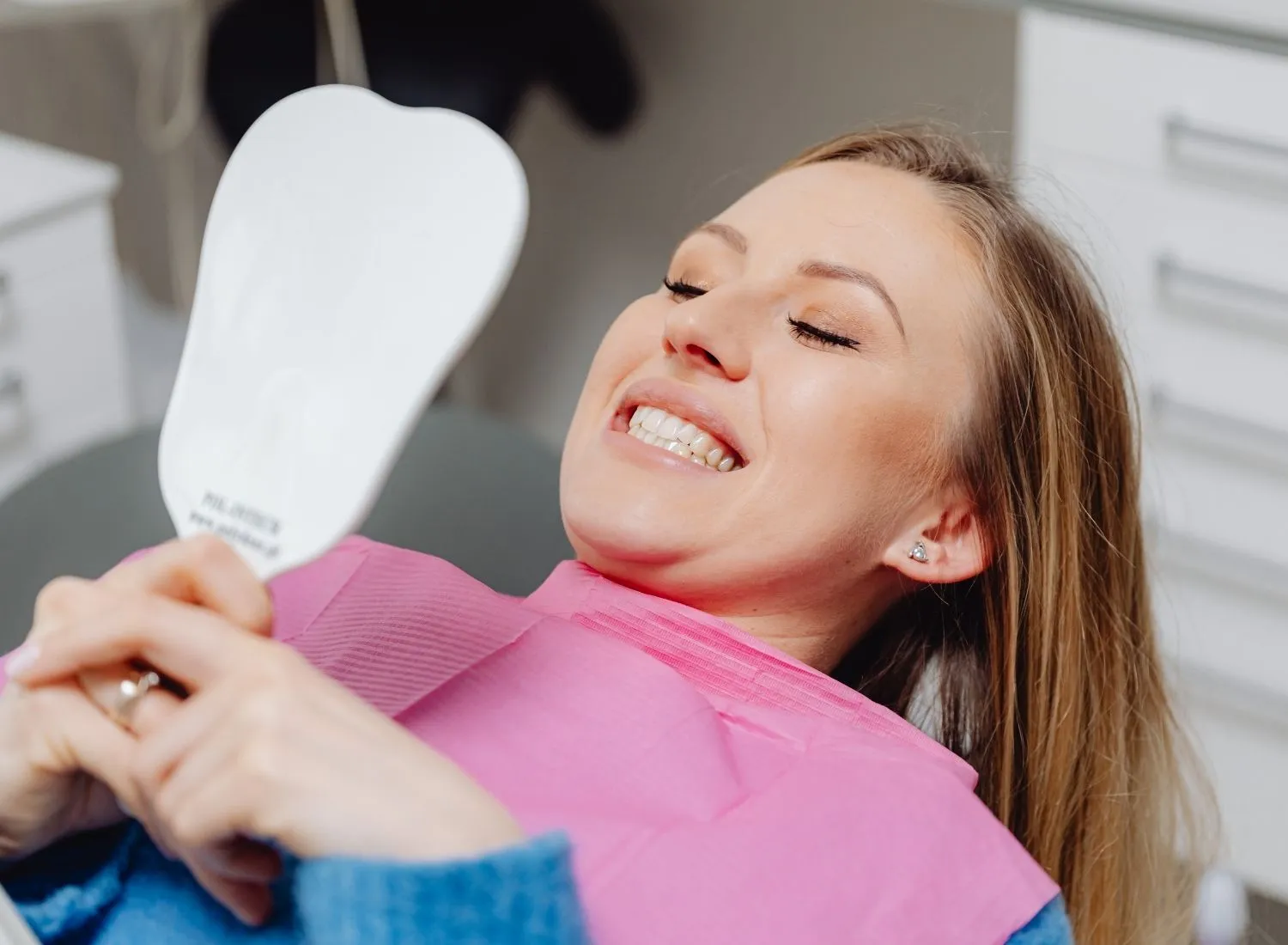 Woman in dental chair wearing pink bib smiles while holding a mirror reflecting her teeth.
