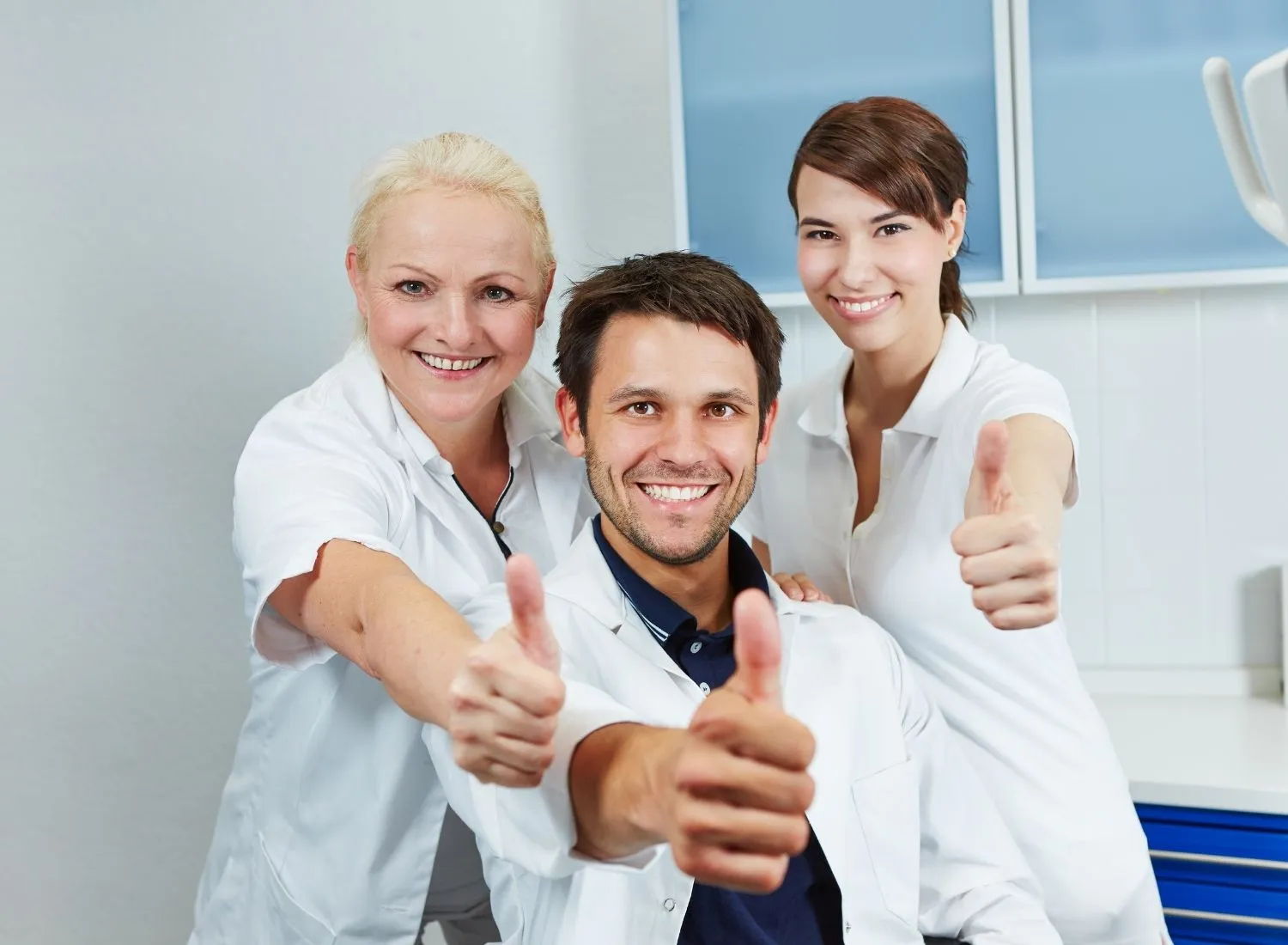 Three smiling medical professionals giving thumbs up in a clinical setting.