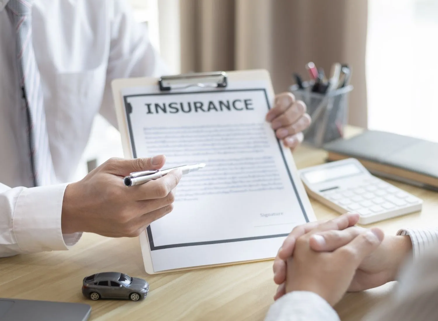 Person showing an insurance document on a clipboard with a pen, with another person's hands folded on table.