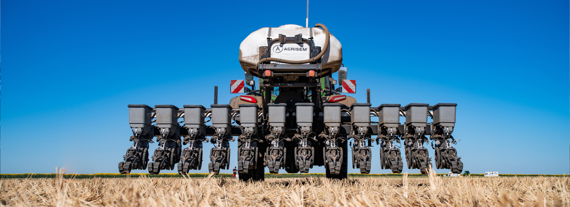 Echipament agricol modern pentru semănat, văzut din față în câmp deschis pe fundalul cerului albastru clar.