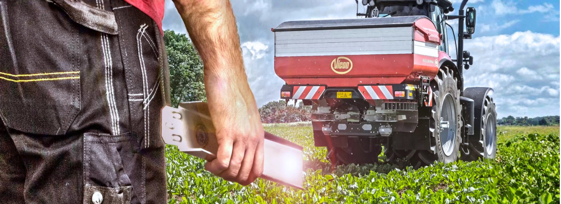 Un fermier în pantaloni scurți negri ține o tabletă în mână, privind un tractor roșu dotat cu un distribuitor de îngrășăminte care traversează un câmp verde sub un cer înnorat.