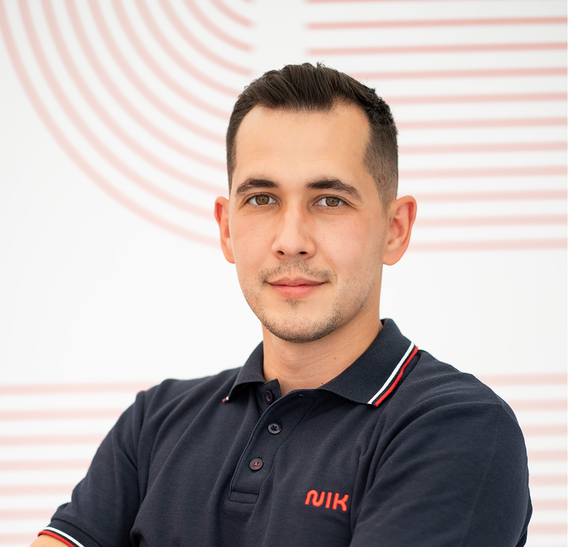 Bărbat tânăr cu păr scurt și castaniu, cu privire calmă, purtând un tricou polo bleumarin cu detalii roșii și albe și un logo roșu pe piept, pozând pe un fundal alb cu linii curbe roșiatice.
