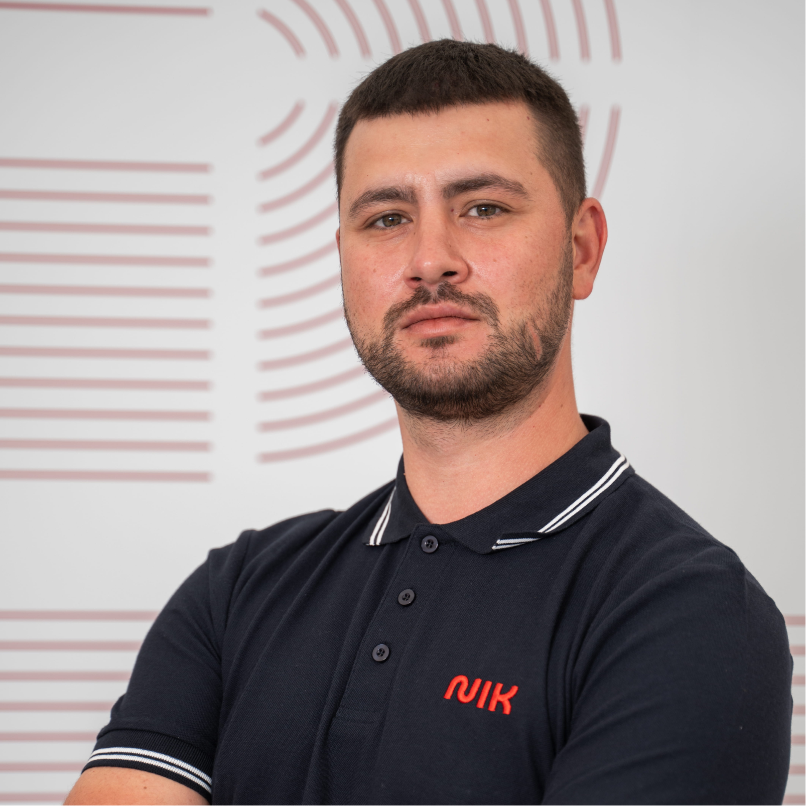 Bărbat cu păr scurt și barbă scurtă, purtând un tricou polo bleumarin cu dungi albe la guler și pe mâneci, cu logo roșu pe piept, în fața unui perete alb cu linii roz deschis.