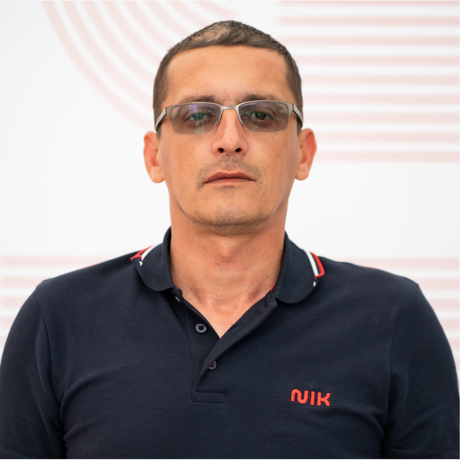 Bărbat cu păr scurt și castaniu, purtând ochelari cu ramă argintie și cămașă polo bleumarin cu logo roșu "NIK", pozând într-un cadru cu fundal alb cu linii curbate roșii.