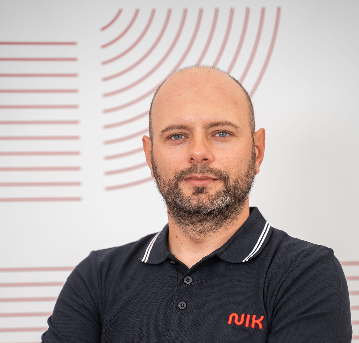 Bărbat cu păr blond subțire și barbă, purtând un tricou negru cu guler și inscripția roșie "NIK" pe piept, pozând cu brațele încrucișate în fața unui perete alb cu design grafic roșu.