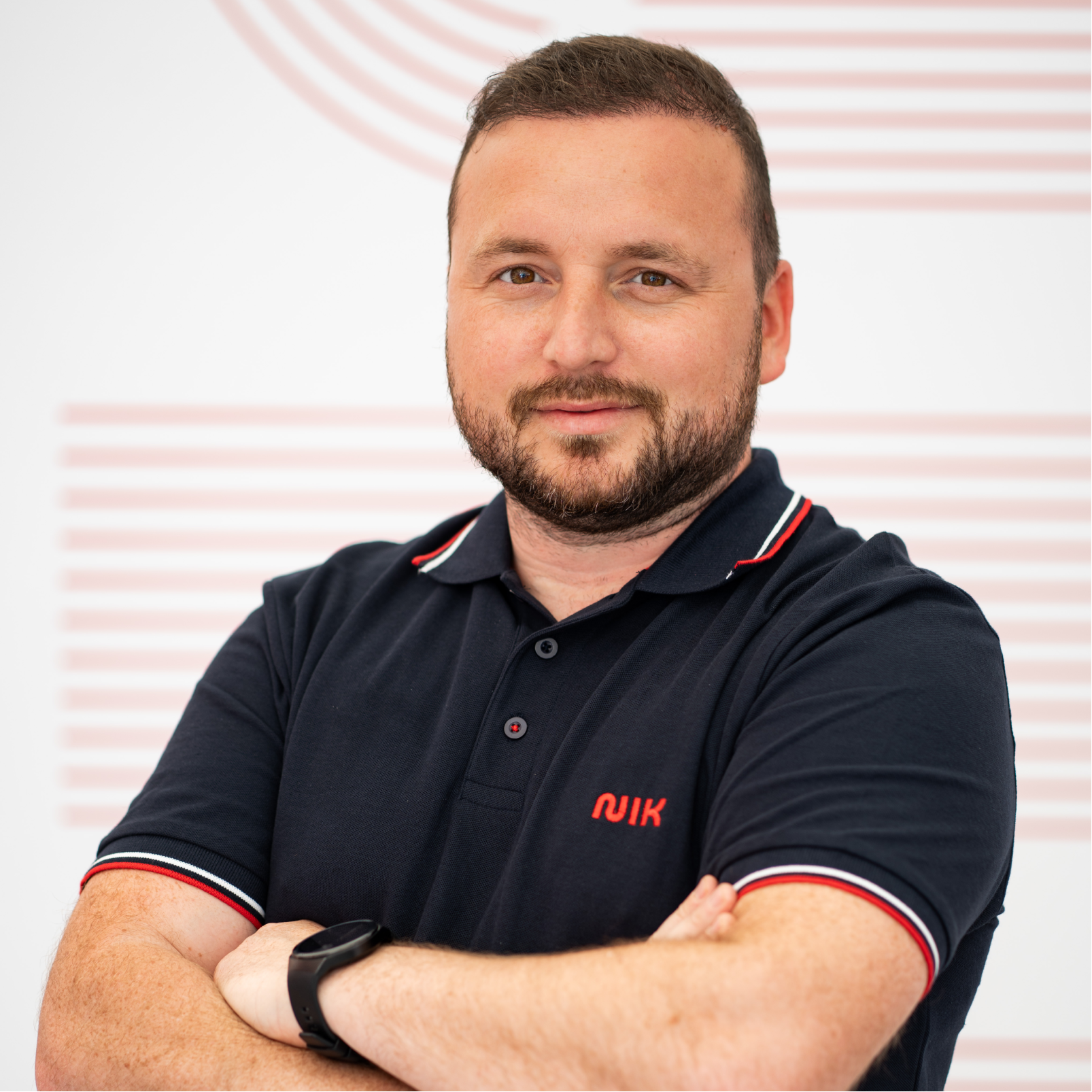 Bărbat cu barbă și păr scurt, îmbrăcat într-un tricou polo bleumarin cu detalii roșii și albe la guler și mâneci, cu brațele încrucișate, purtând un ceas negru, pe fundal alb cu linii orizontale roșii.
