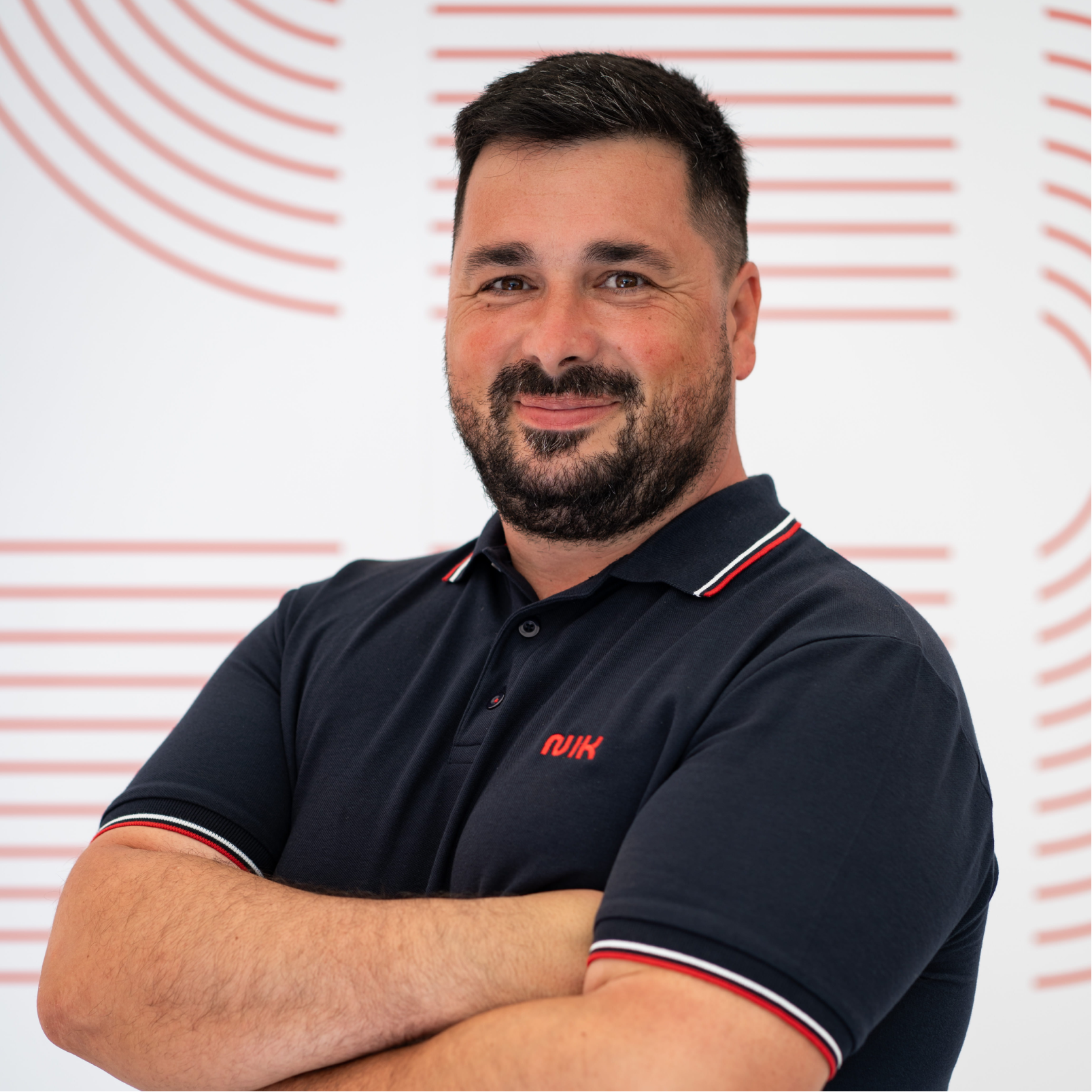 Bărbat cu păr și barbă scurtă, zâmbind ușor, purtând tricou polo bleumarin cu dungi roșii și albe la guler și mâneci, stând cu brațele încrucișate în fața unui perete alb cu modele roșii curbe și drepte.