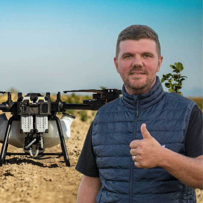 Bărbat cu veste albastră în câmp arid, dând un semn de apreciere cu degetul mare, lângă un drone agricol cu elice pe fundalul cerului senin.