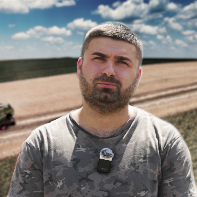 Bărbat cu păr scurt și barbă purtând un tricou gri camuflaj, cu microfon agățat de gât, stând într-un câmp agricol cu cereale coapte și un combină de recoltat pe fundal sub un cer albastru cu nori.