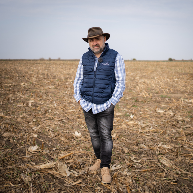 Bărbat cu pălărie maro, vestă albastră fără mâneci, cămașă în carouri albă și albastră și pantaloni negri stând într-un câmp uscat cu resturi de porumb pe sol sub cer senin.