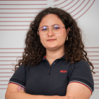 Tânără cu păr creț, purtând ochelari rotunzi și o bluză polo neagră cu logo roșu, stând cu brațele încrucișate în fața unui perete alb cu linii roșii paralele curbate.