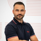 Bărbat cu păr și barbă scurtă, purtând un tricou polo negru cu detalii roșii, pozând cu brațele încrucișate și zâmbind subtil pe un fundal deschis cu linii curbe roșii și gri.