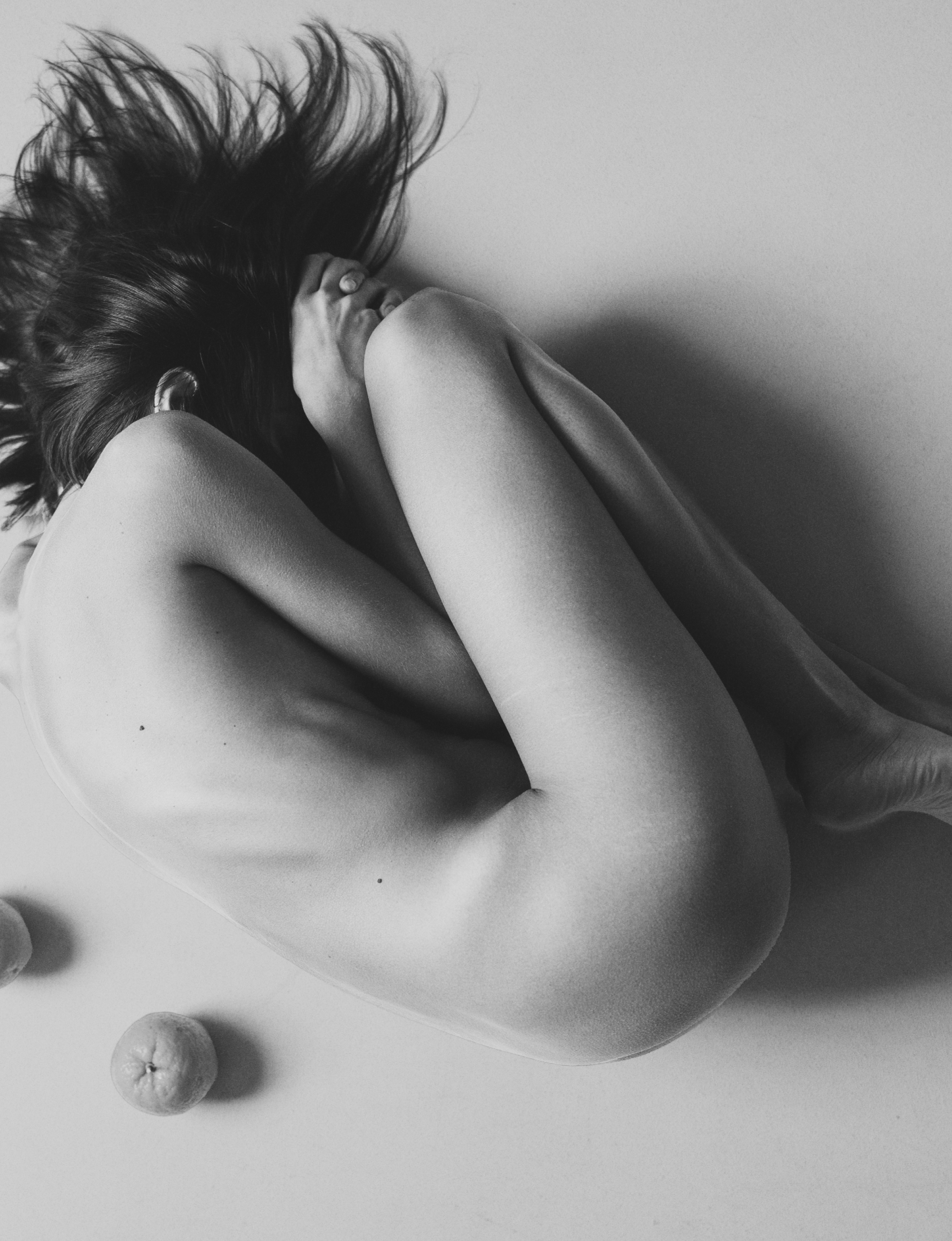 Black-and-white editorial photograph of a curled nude figure surrounded by scattered oranges.