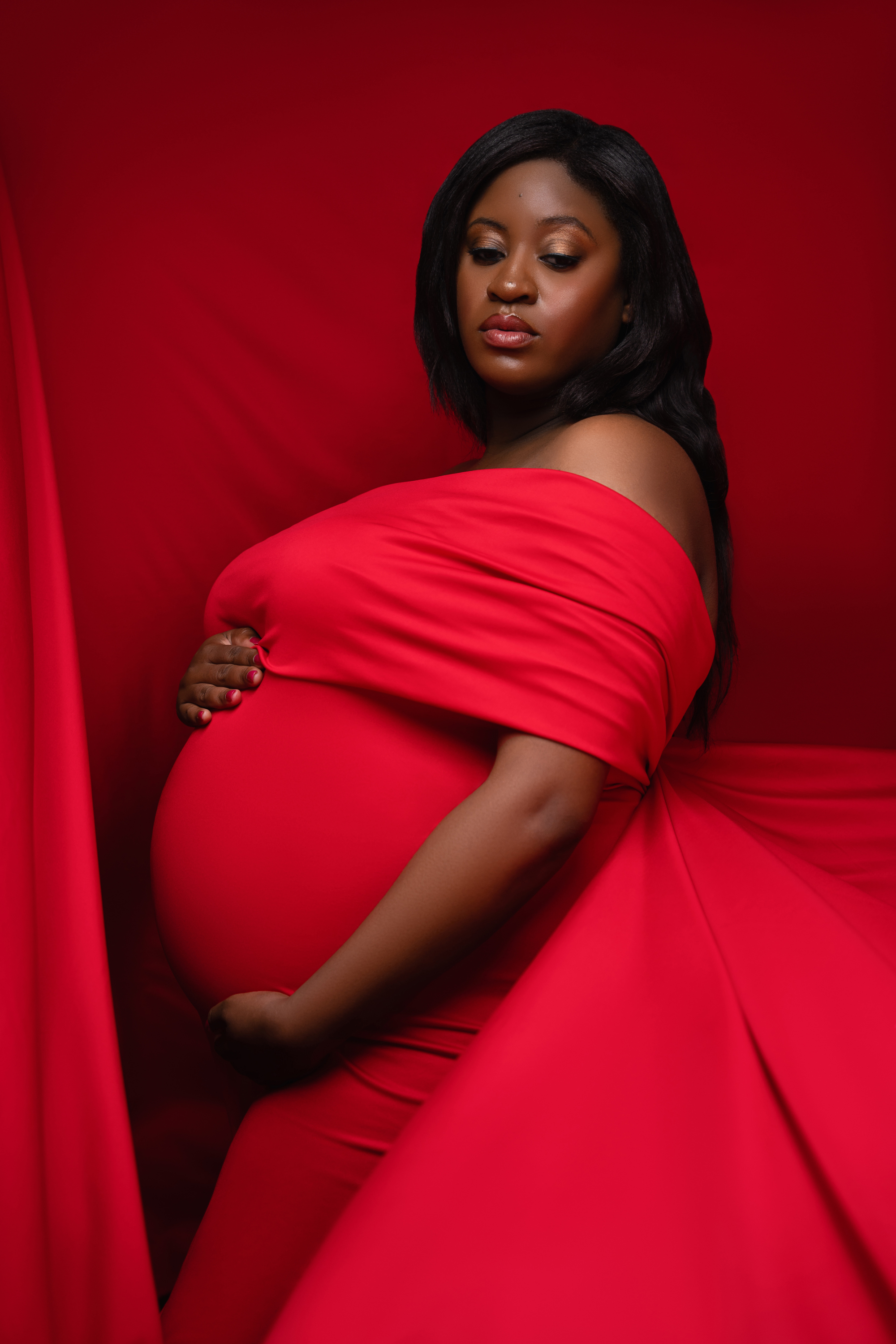 Pregnant woman draped in a flowing red gown against a dark studio background.