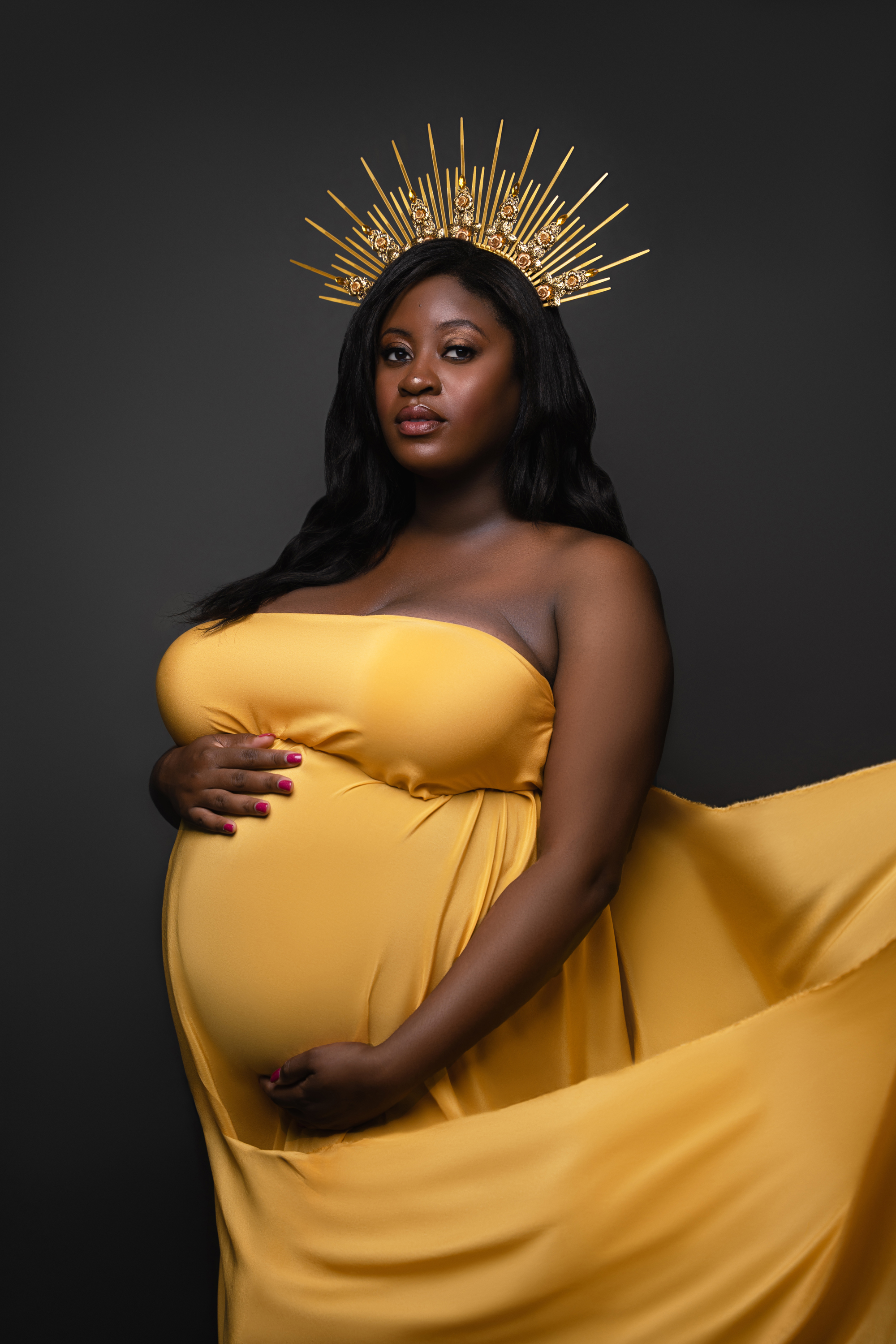 Pregnant woman wrapped in a sculptural gold gown against a dark studio background.