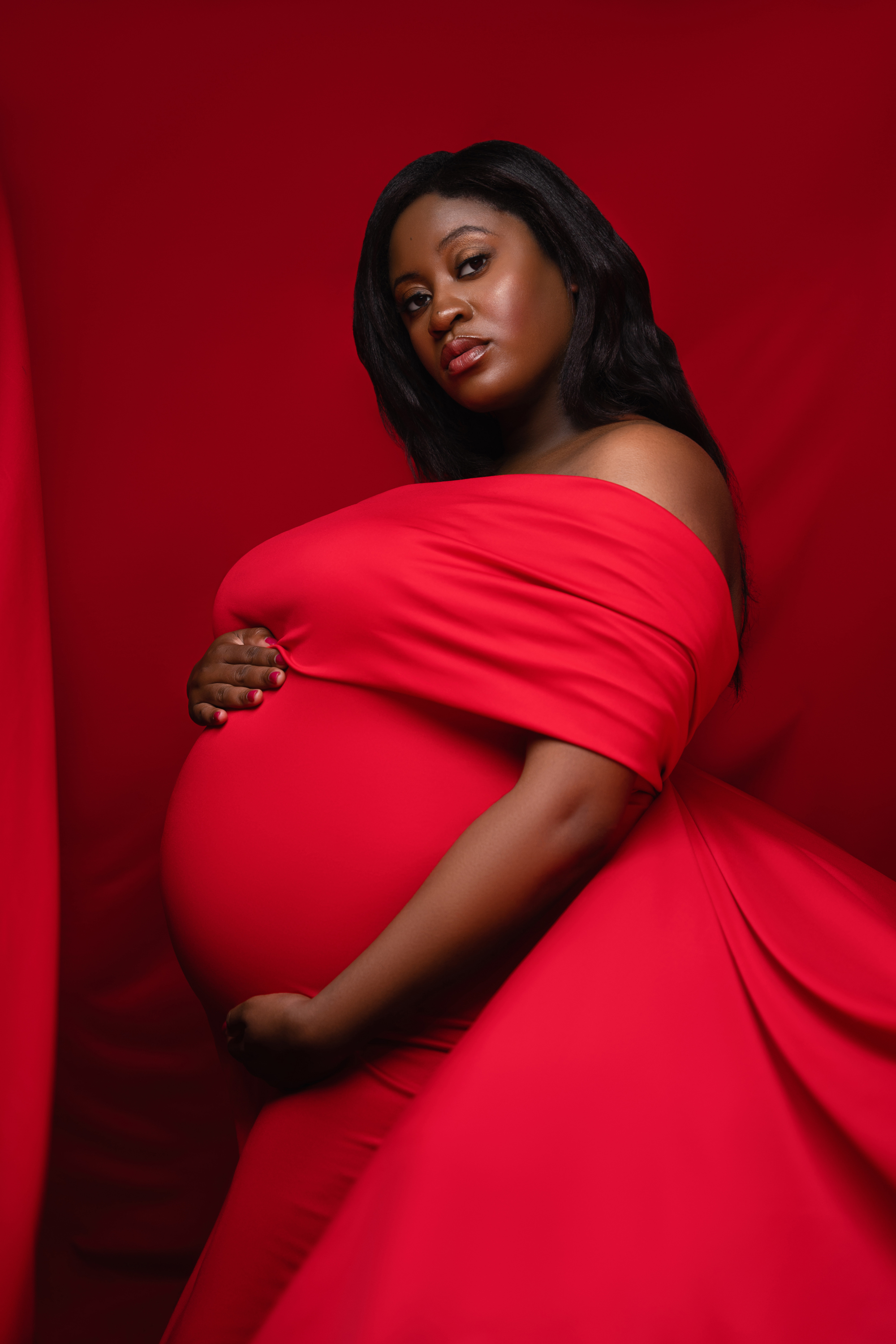 Maternity portrait of a woman in a flowing red gown with dramatic studio lighting.