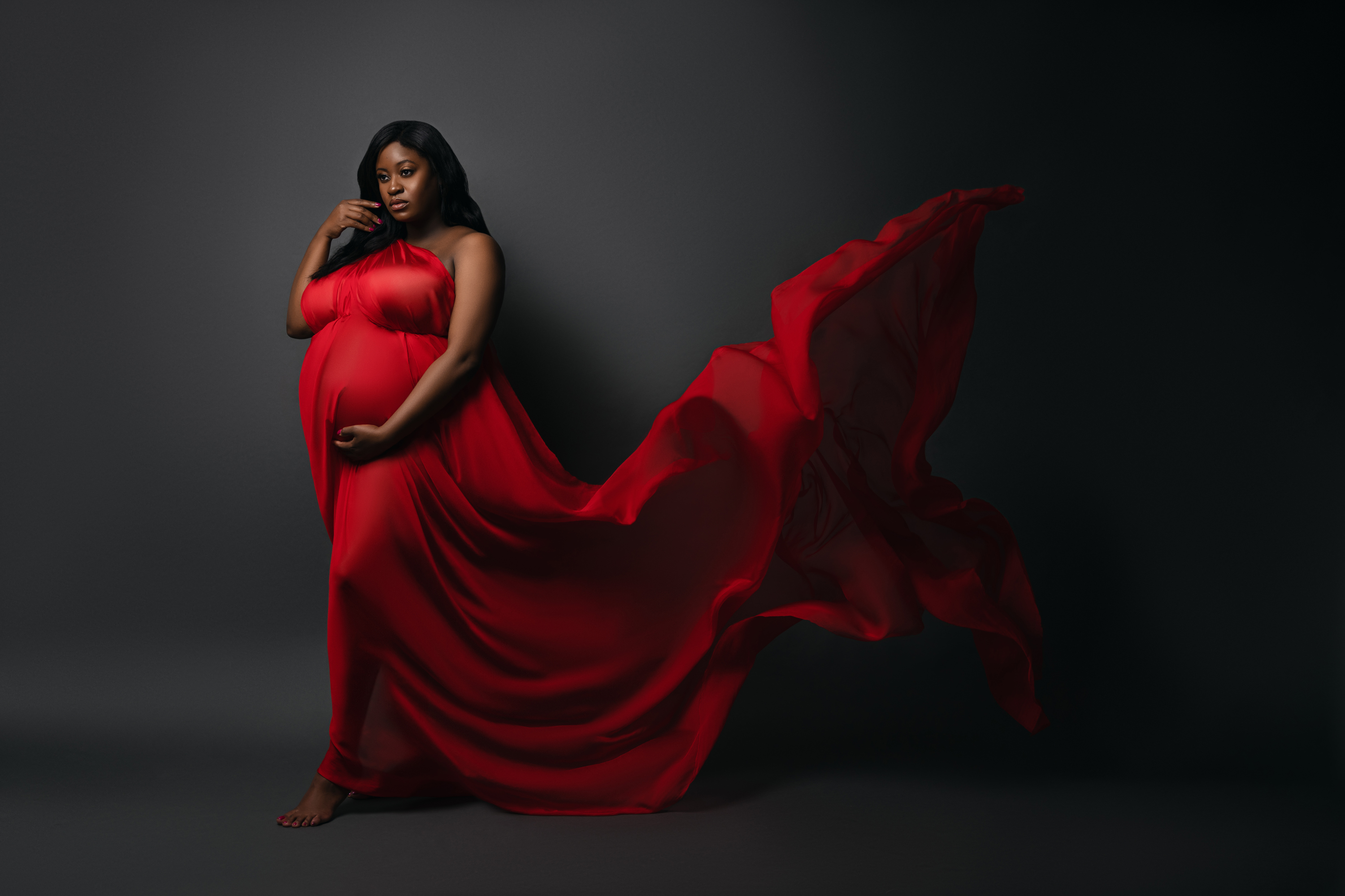 Dramatic maternity portrait in a flowing red gown with fabric extending across a dark studio setting.