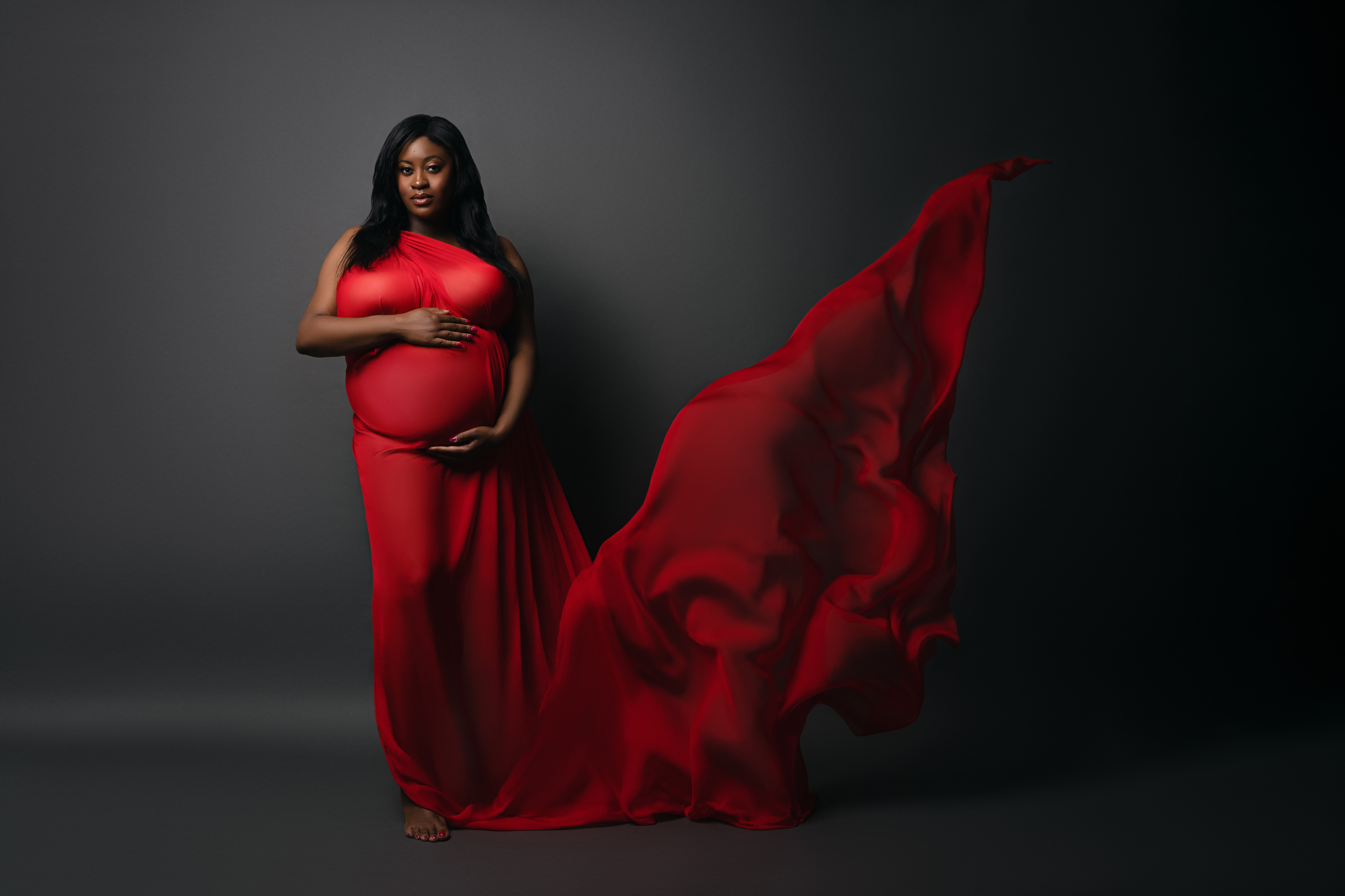 Pregnant woman in a red gown posed against a dark studio background with flowing fabric.
