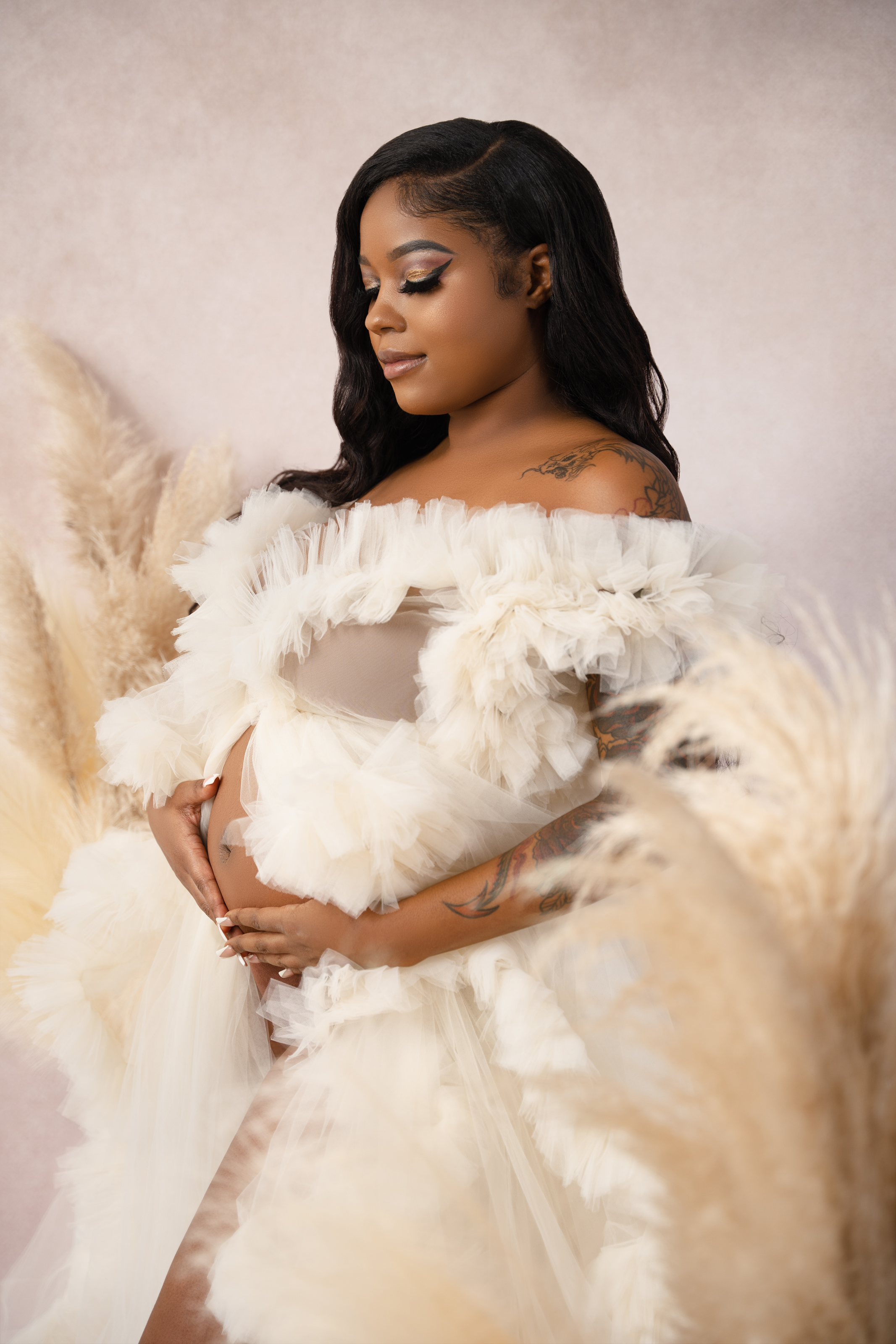 Close maternity portrait of a woman in a cream feathered gown against a soft blush studio background.
