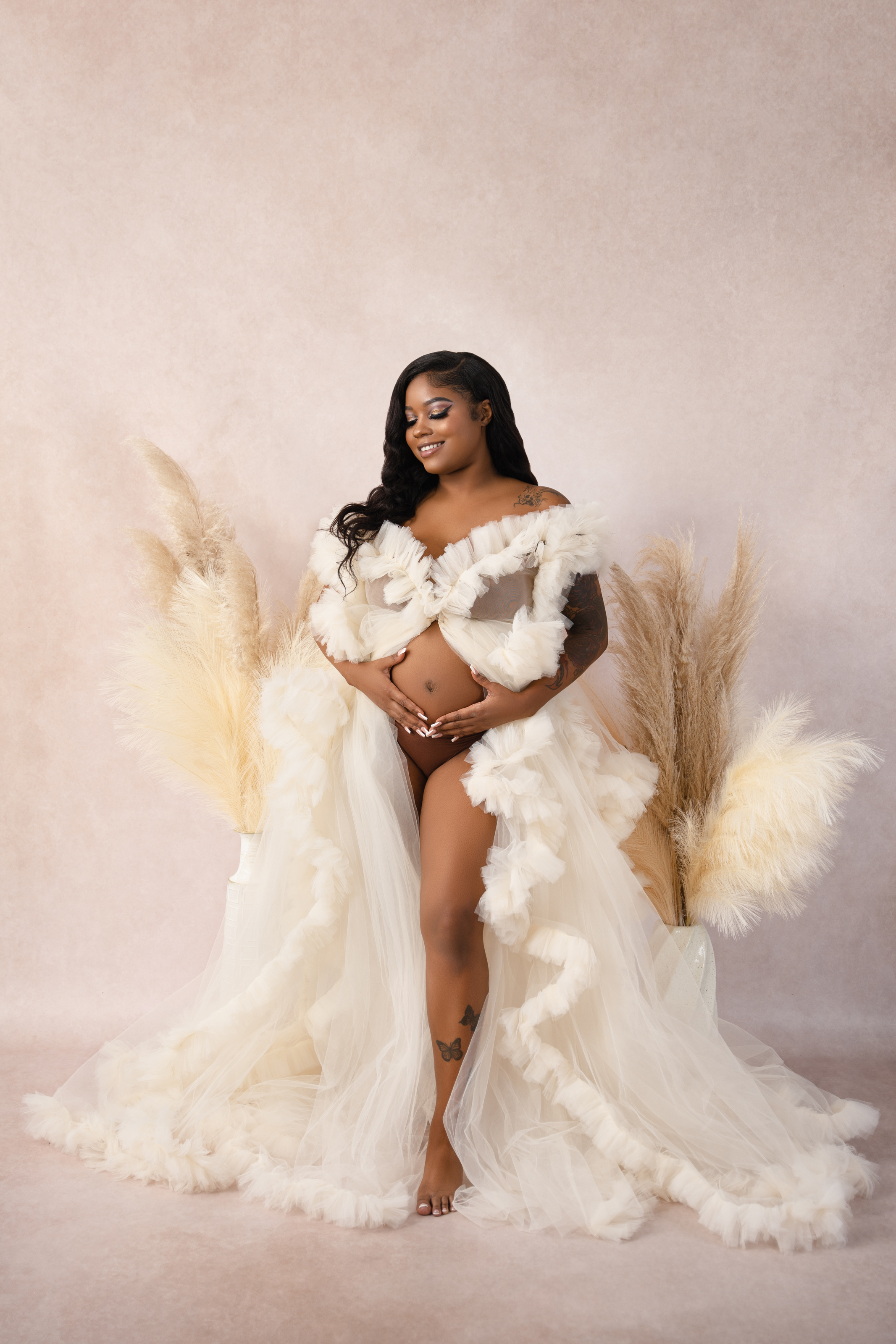 Full-length maternity portrait of a woman in a cream feathered gown against a soft blush studio background.