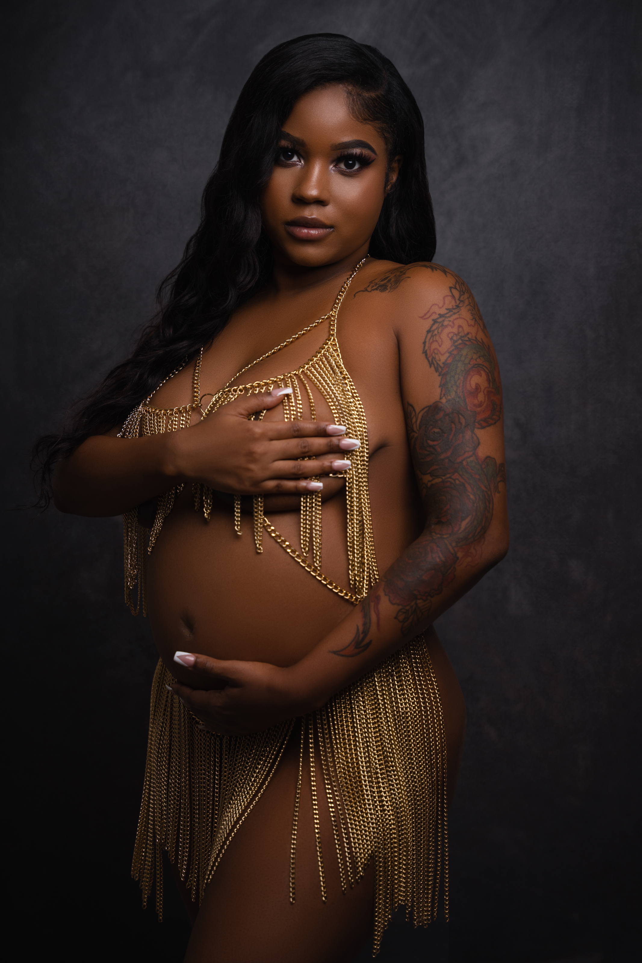 Maternity portrait of a woman in gold fringe styling against a dark textured studio background.