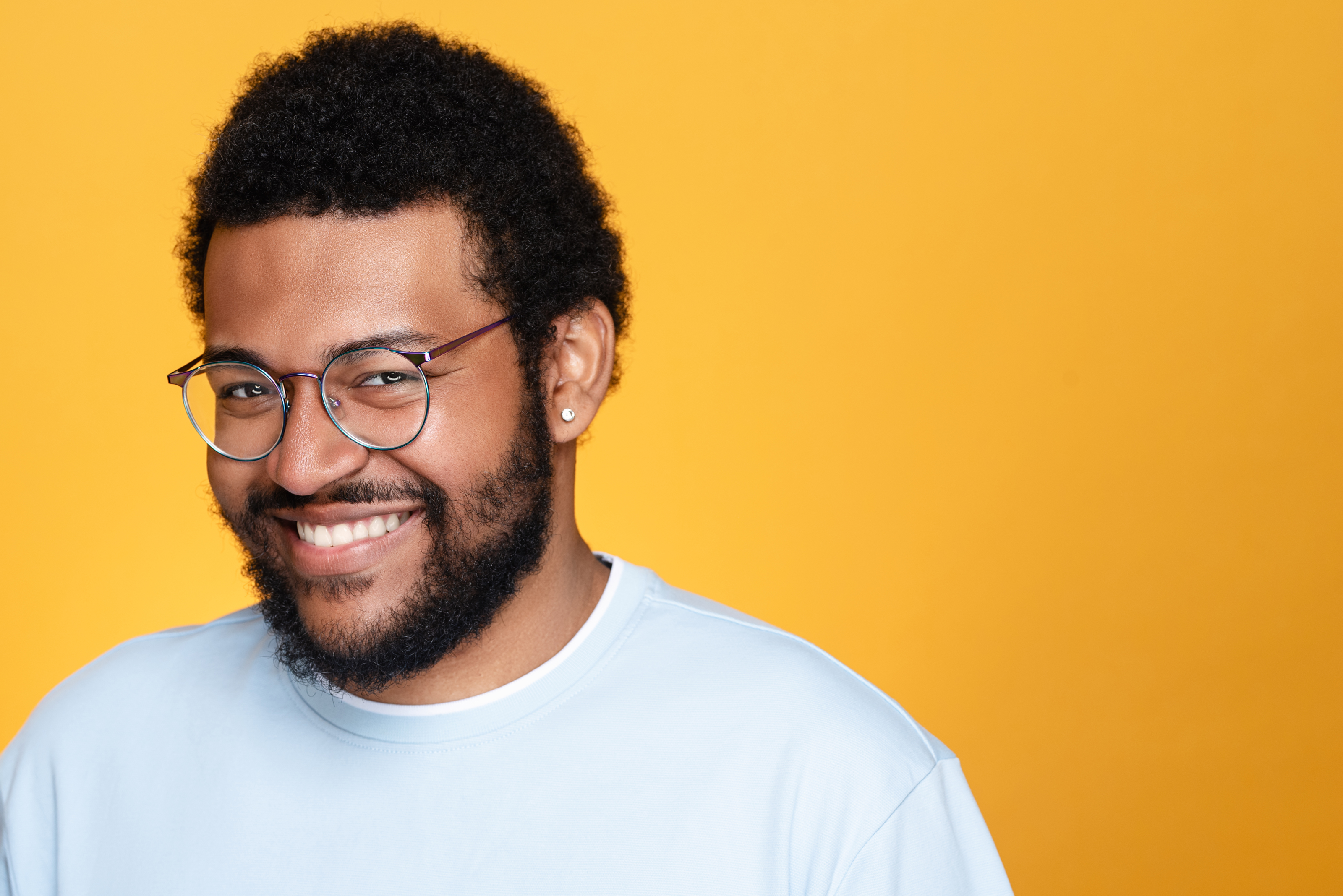 Smiling studio portrait of a man with glasses in a light blue shirt against a bright yellow background.