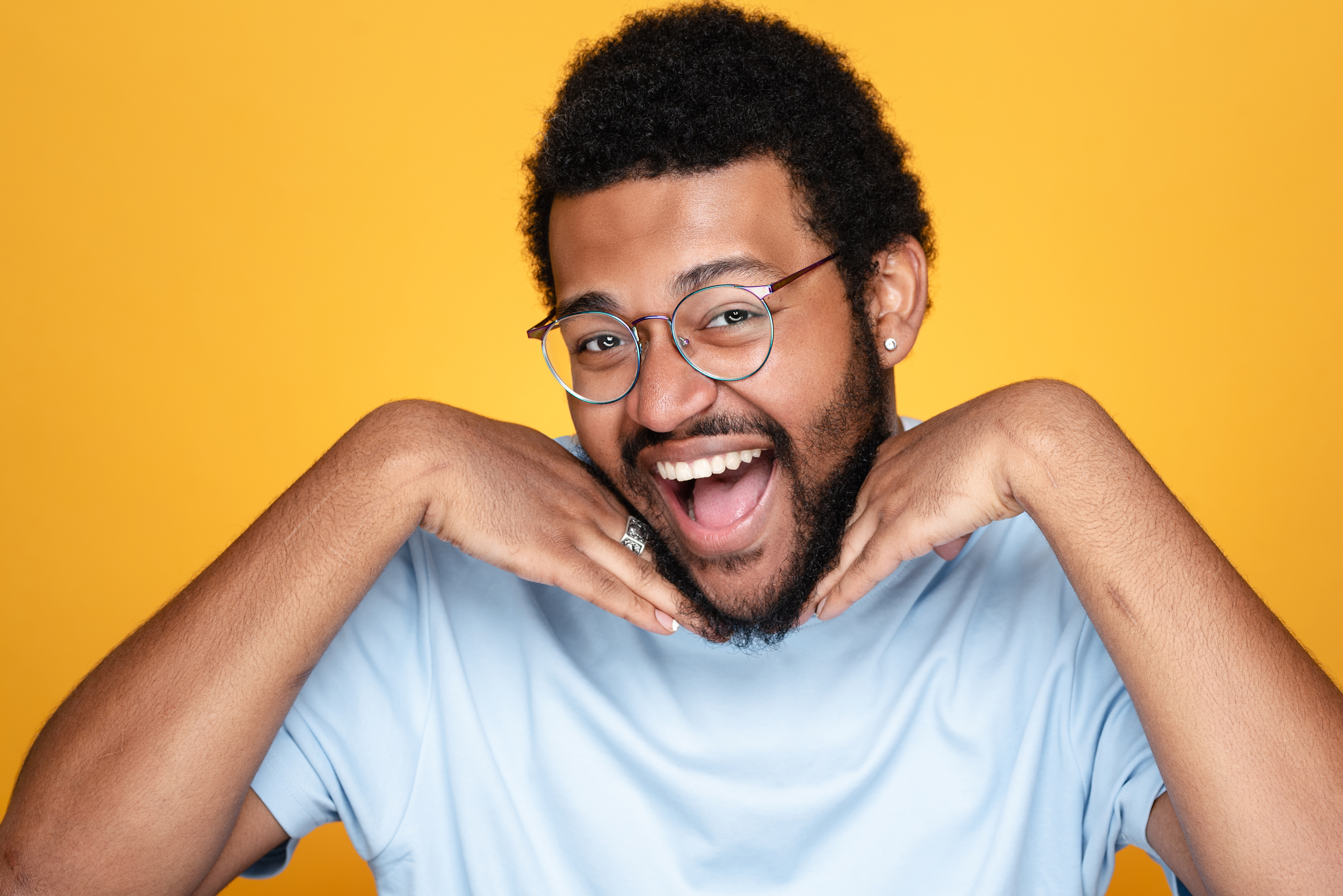 Playful studio portrait of a man posing with his hands under his chin against a bright yellow background.