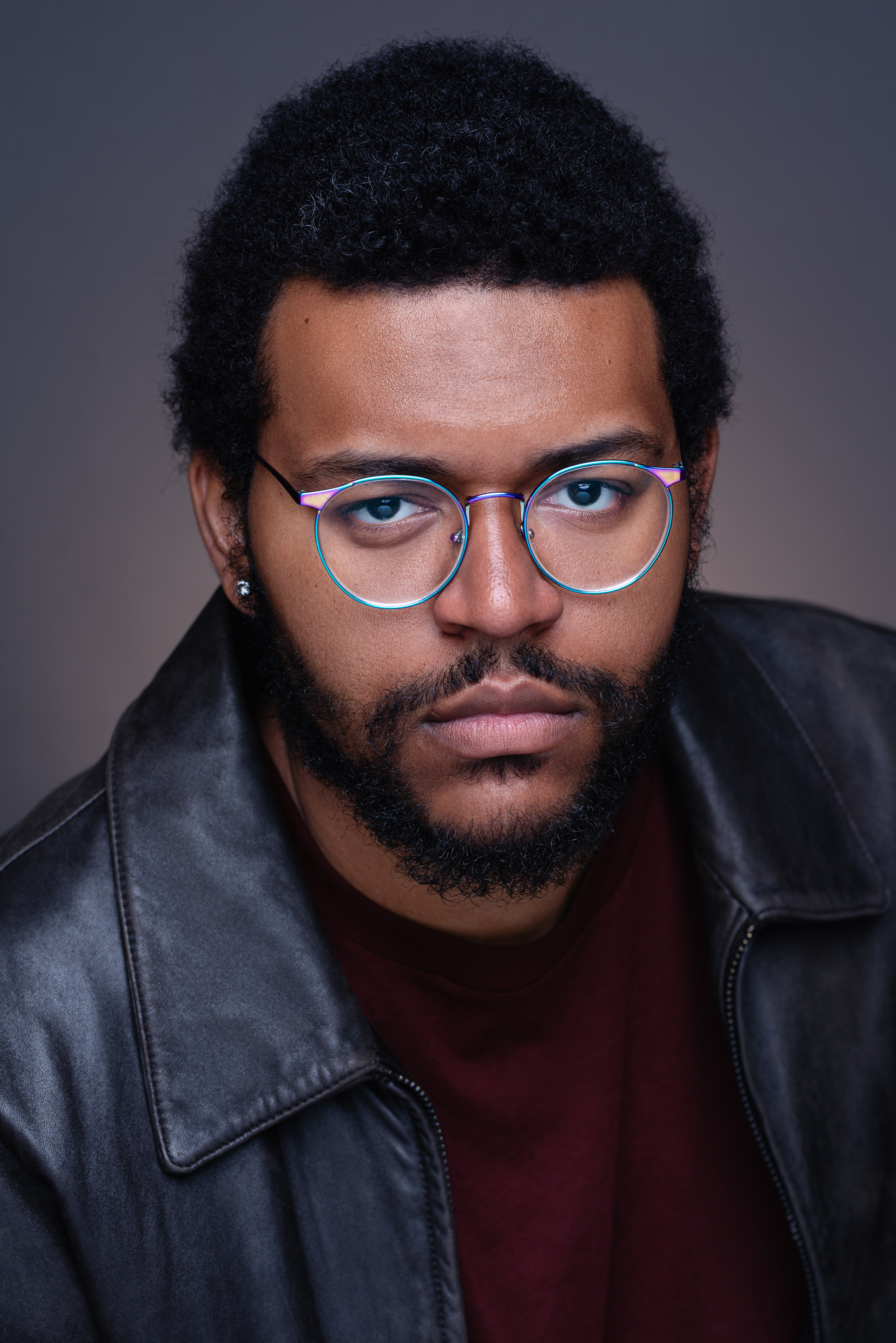 Dramatic studio portrait of a man with glasses wearing a black leather jacket against a dark neutral background.