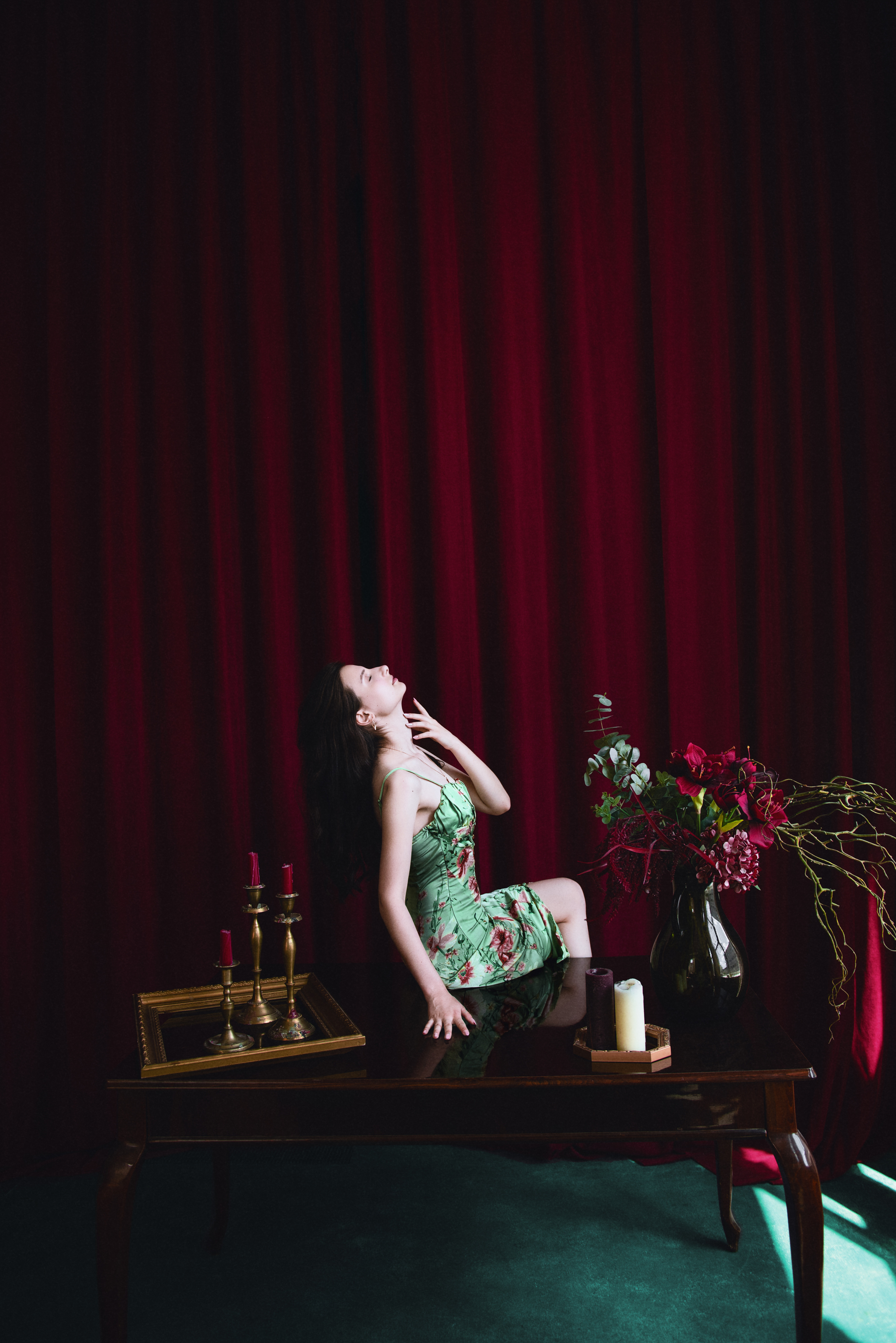 Cinematic portrait of a woman in a floral dress posing on a table against deep red curtains and dramatic studio lighting.