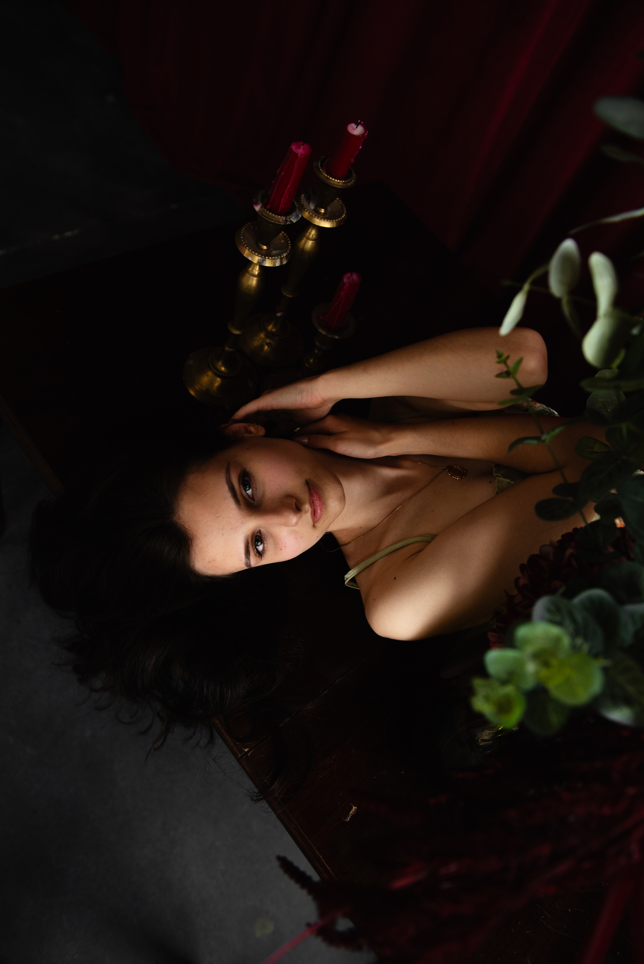 Unretouched overhead portrait of a woman lying among flowers and candleholders in a dark studio setting.