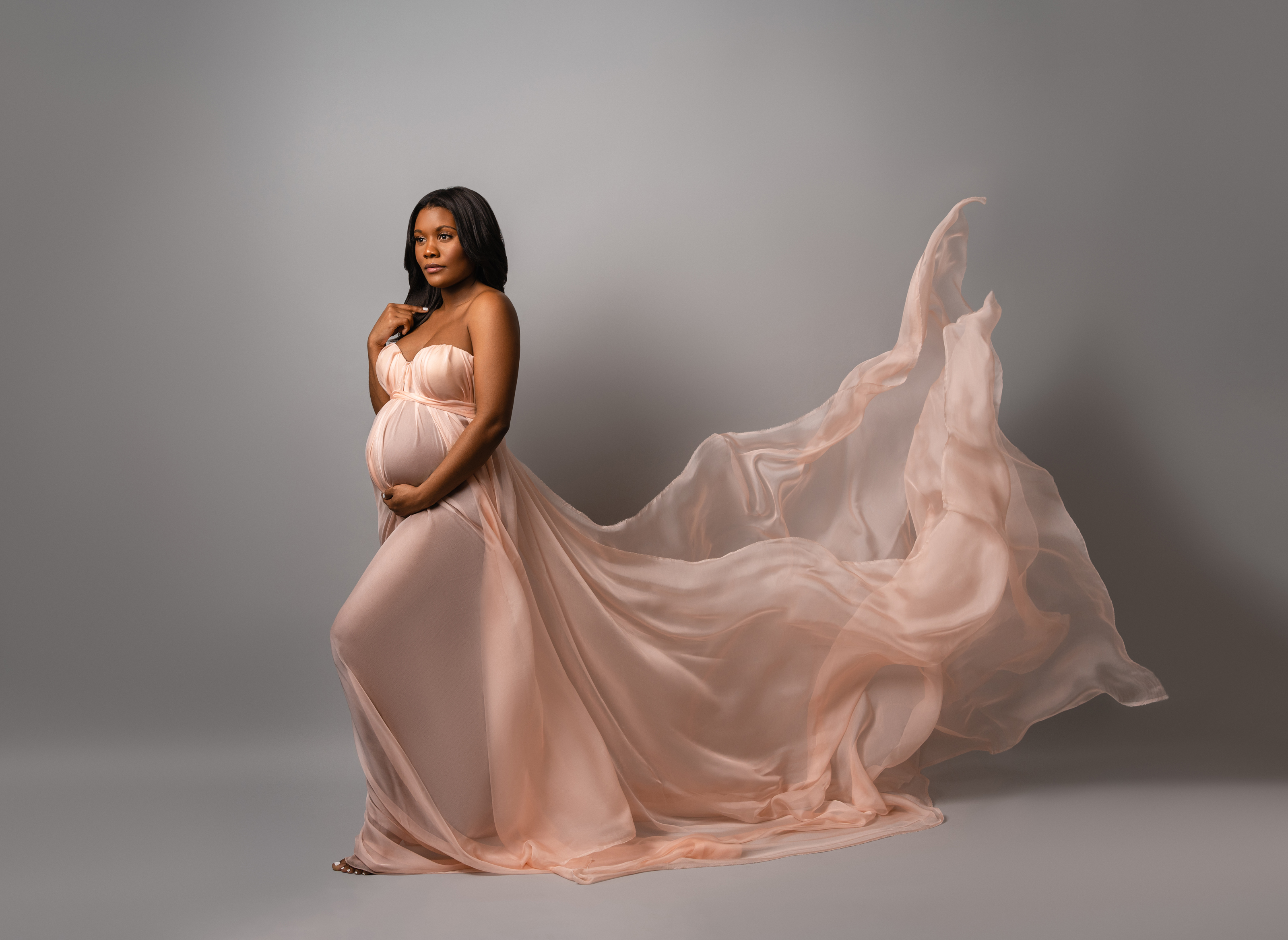 Full-length maternity portrait in a flowing peach gown captured in motion against a neutral studio background.