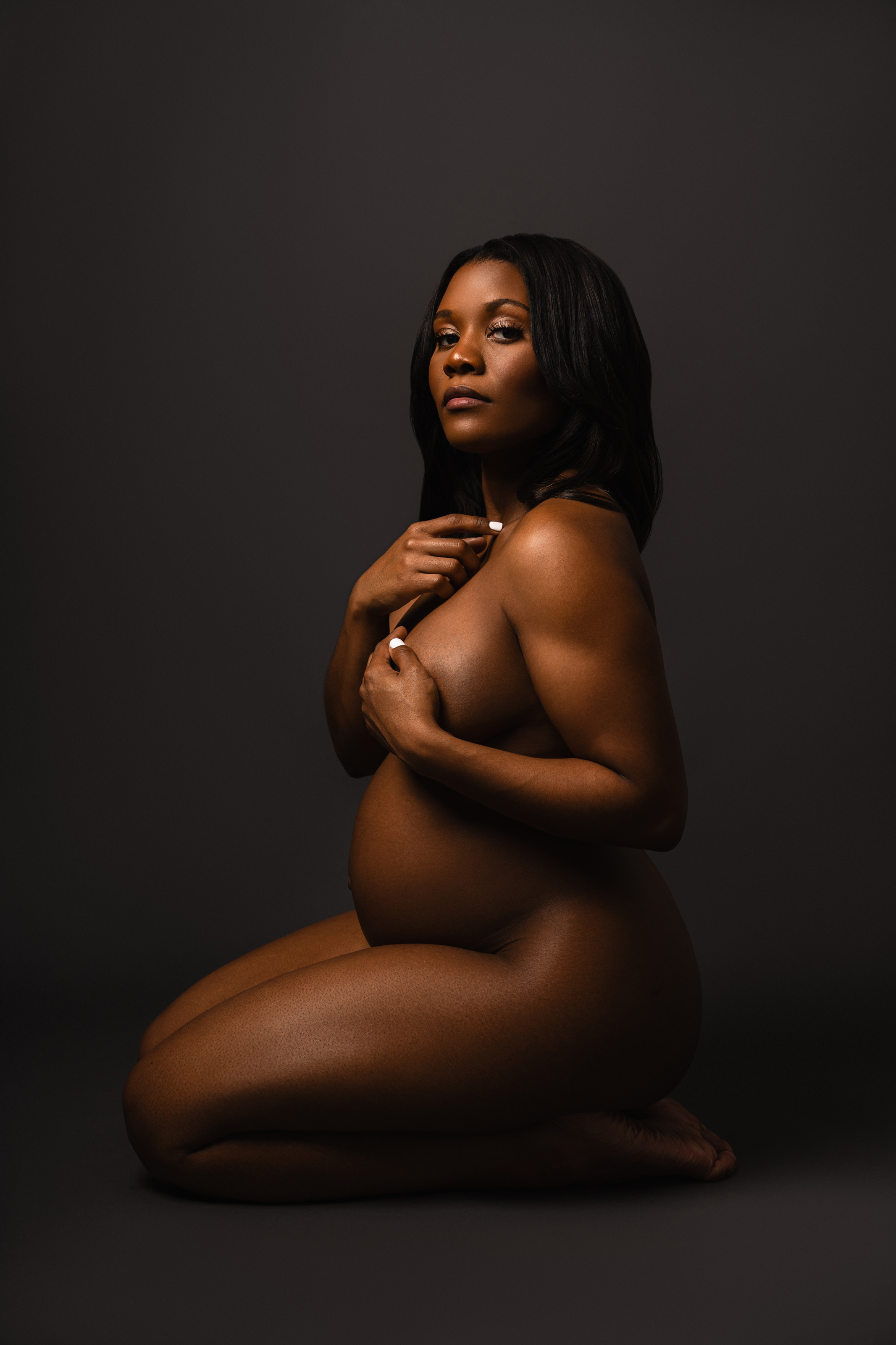 Side maternity portrait of a kneeling pregnant woman photographed with soft low-key studio lighting.