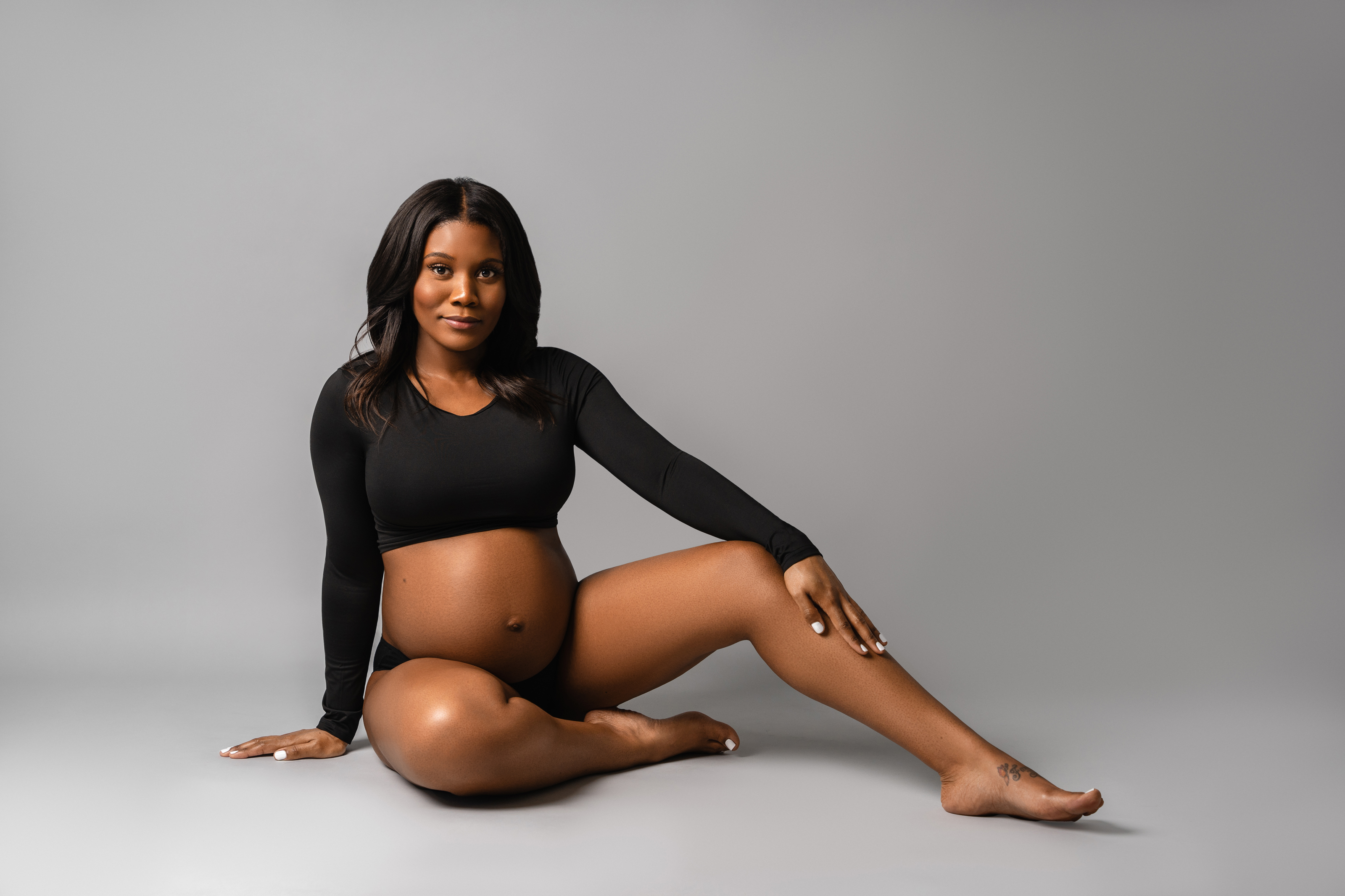 Seated maternity portrait in black styling against a soft gray studio background.