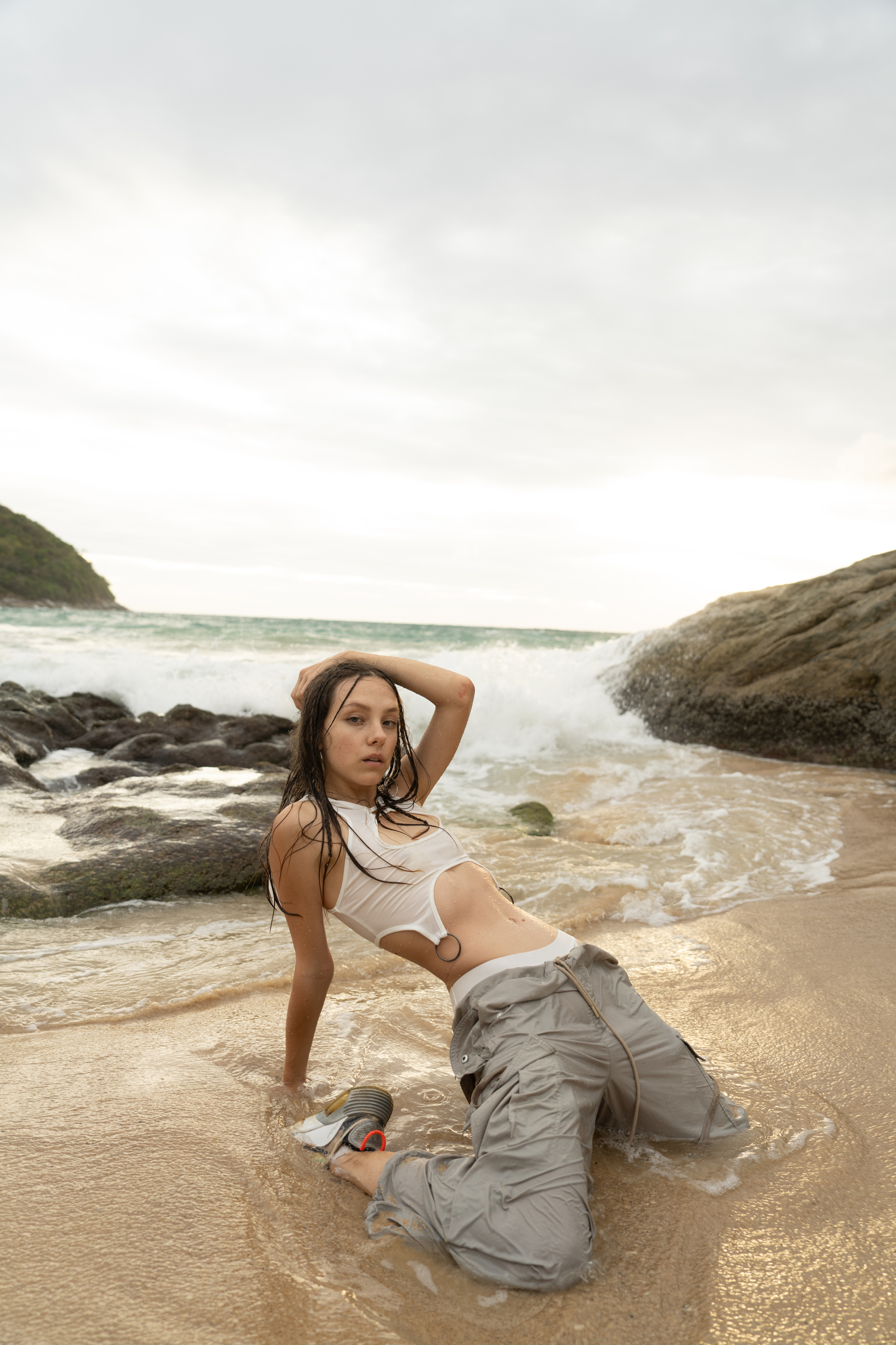 Raw beach portrait of a woman in a white top and cargo trousers posing in shallow water by rocky coastline