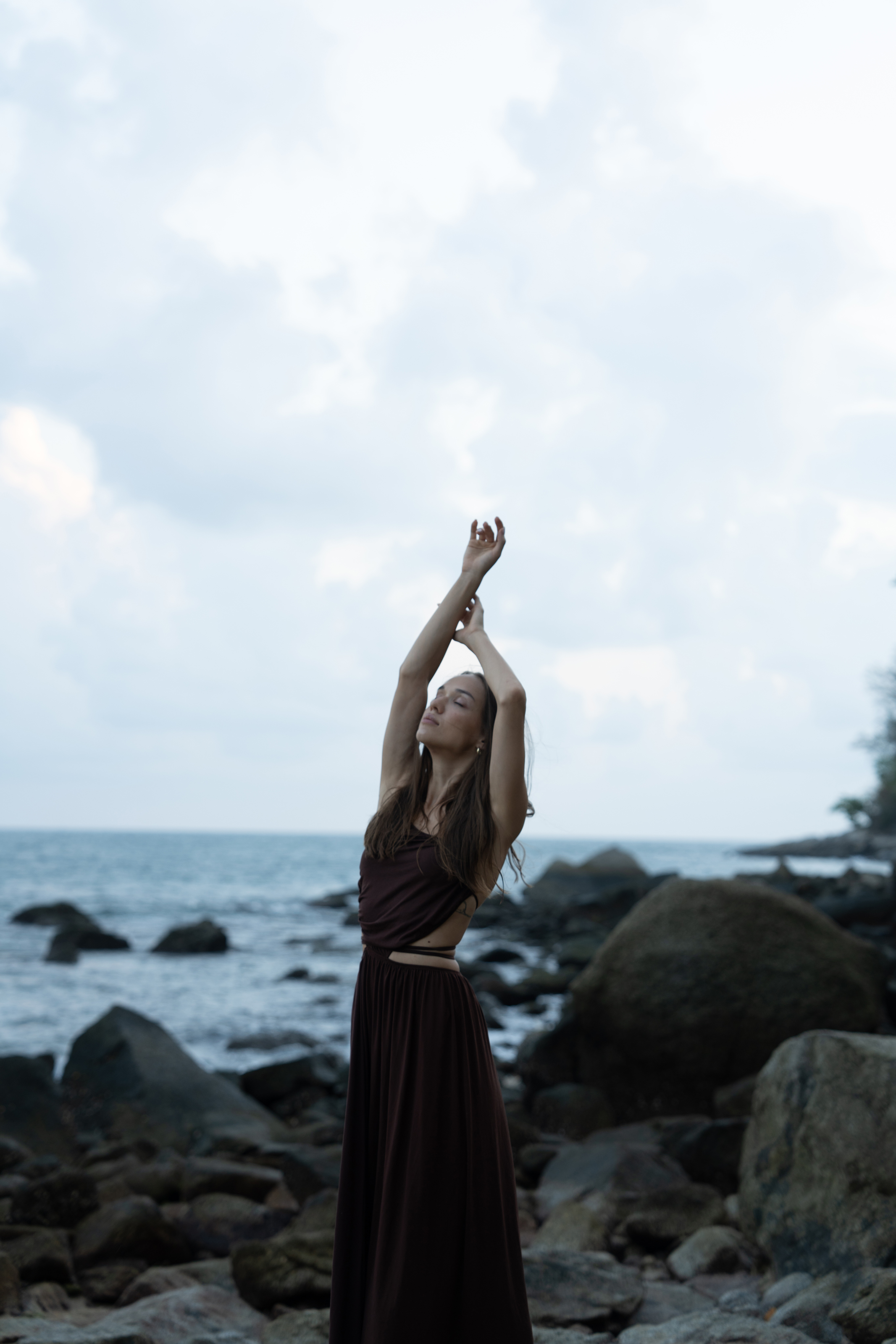 Raw coastal portrait of a woman in a dark flowing dress standing among rocks beside the sea