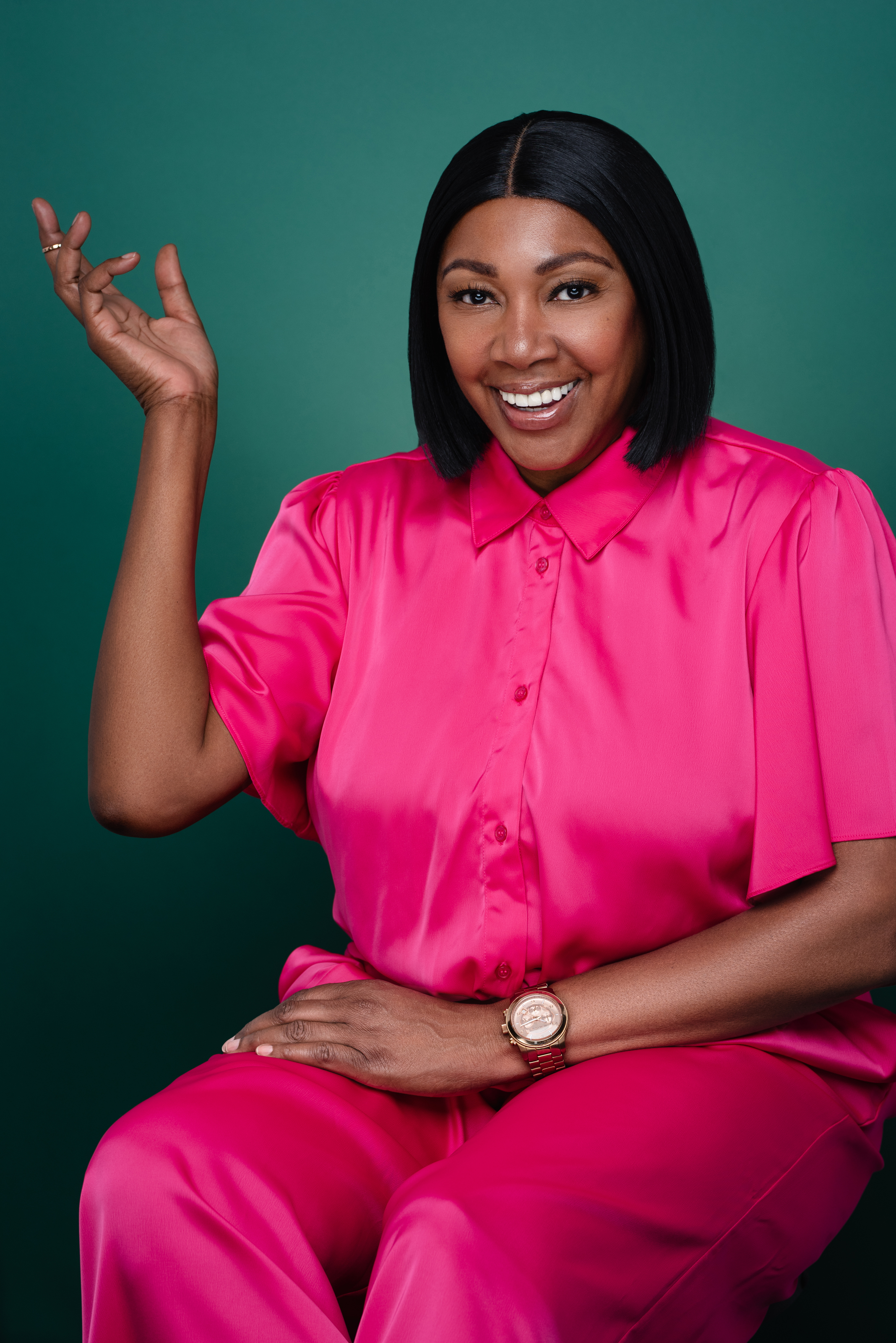 Studio portrait of a smiling woman in a bright pink outfit posing with one hand raised against a green background