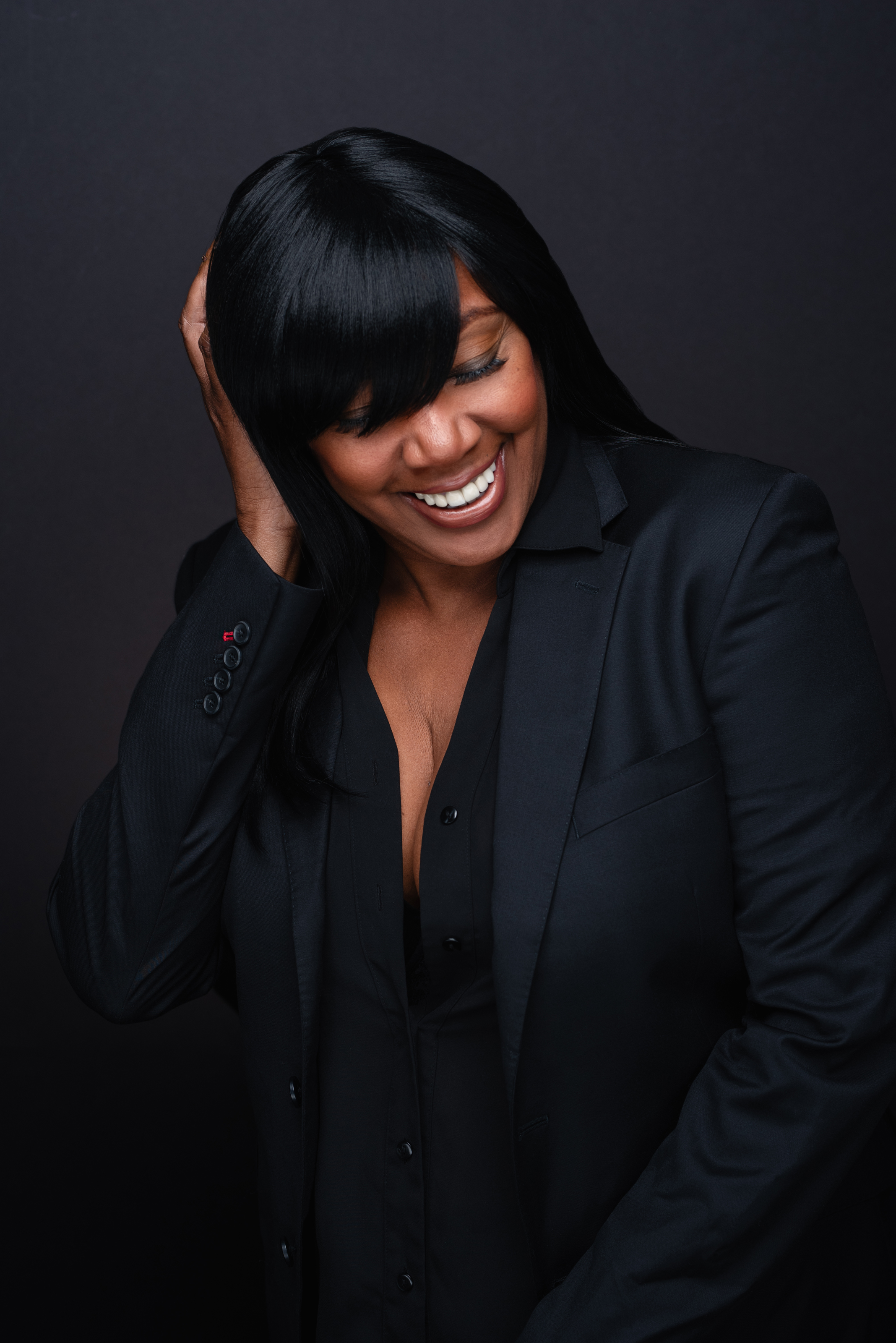 Candid studio portrait of a smiling woman in a black suit with one hand in her hair against a dark background