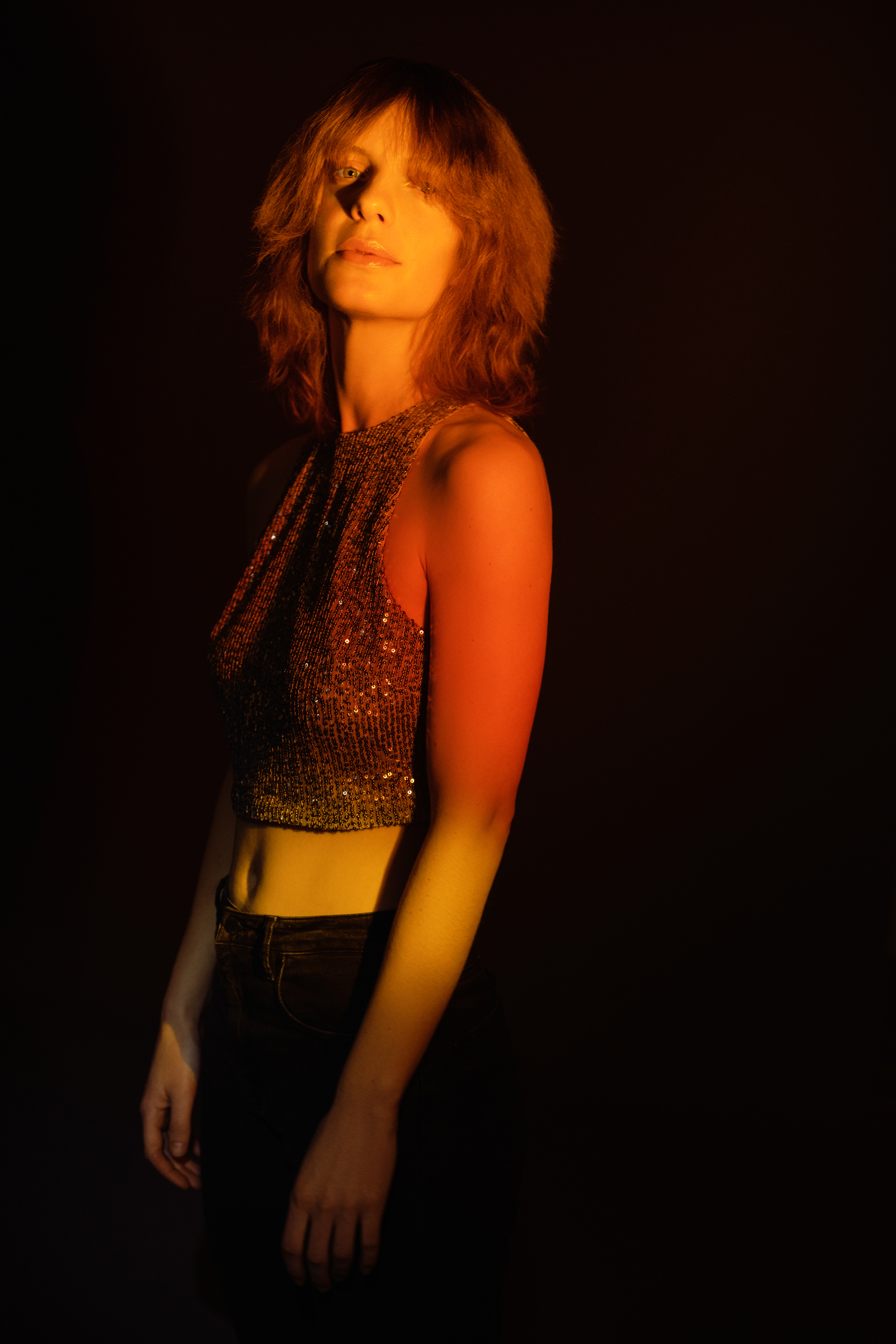 Dramatic studio portrait of a woman in a shimmering top illuminated by warm amber light against a dark background