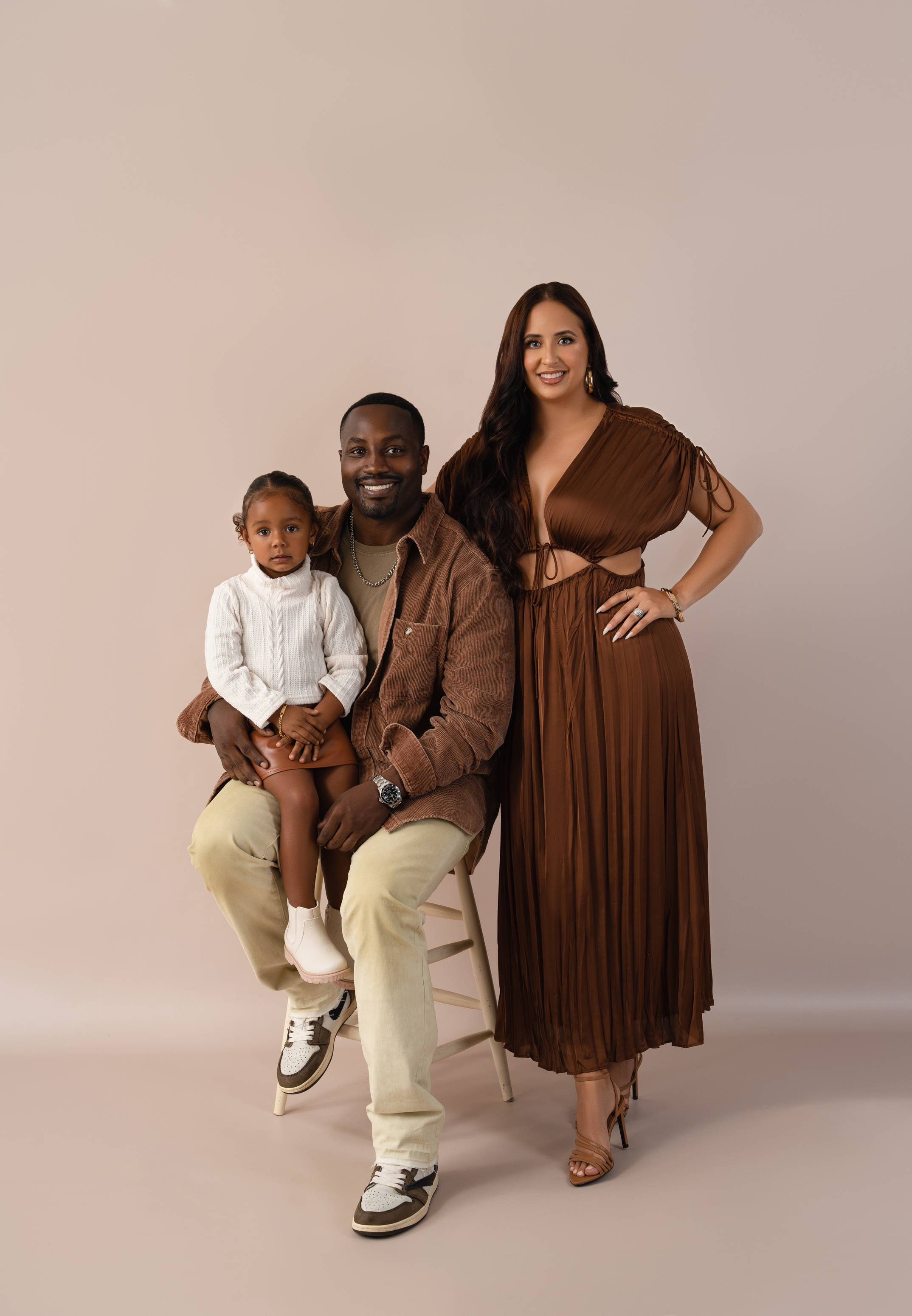 Warm studio family portrait of two parents with their young daughter in coordinated brown and neutral outfits