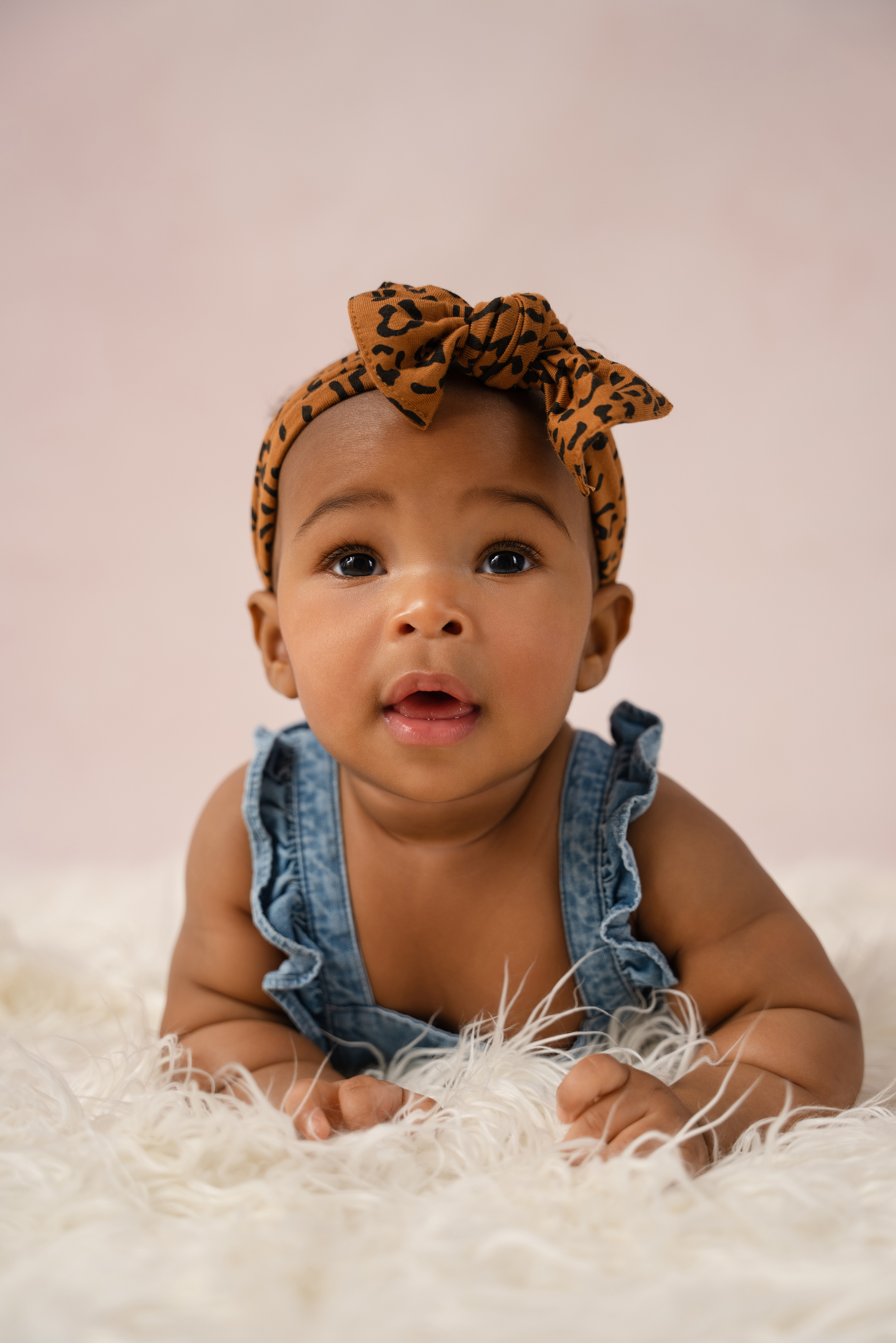 Baby portrait in a blue denim outfit with a patterned headband against a soft pink background