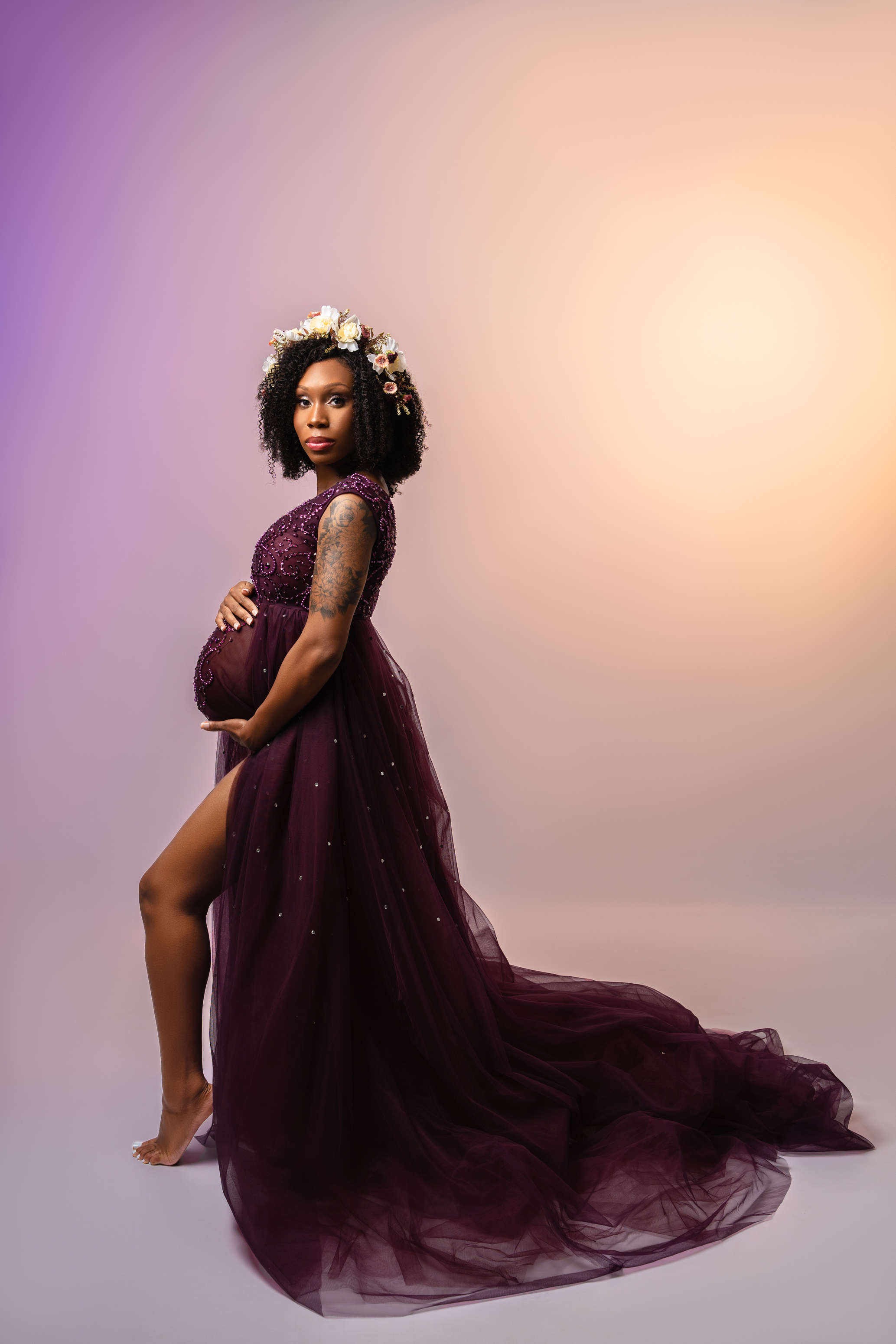 Full-length maternity portrait in a deep plum gown with a long flowing train against a soft gradient background