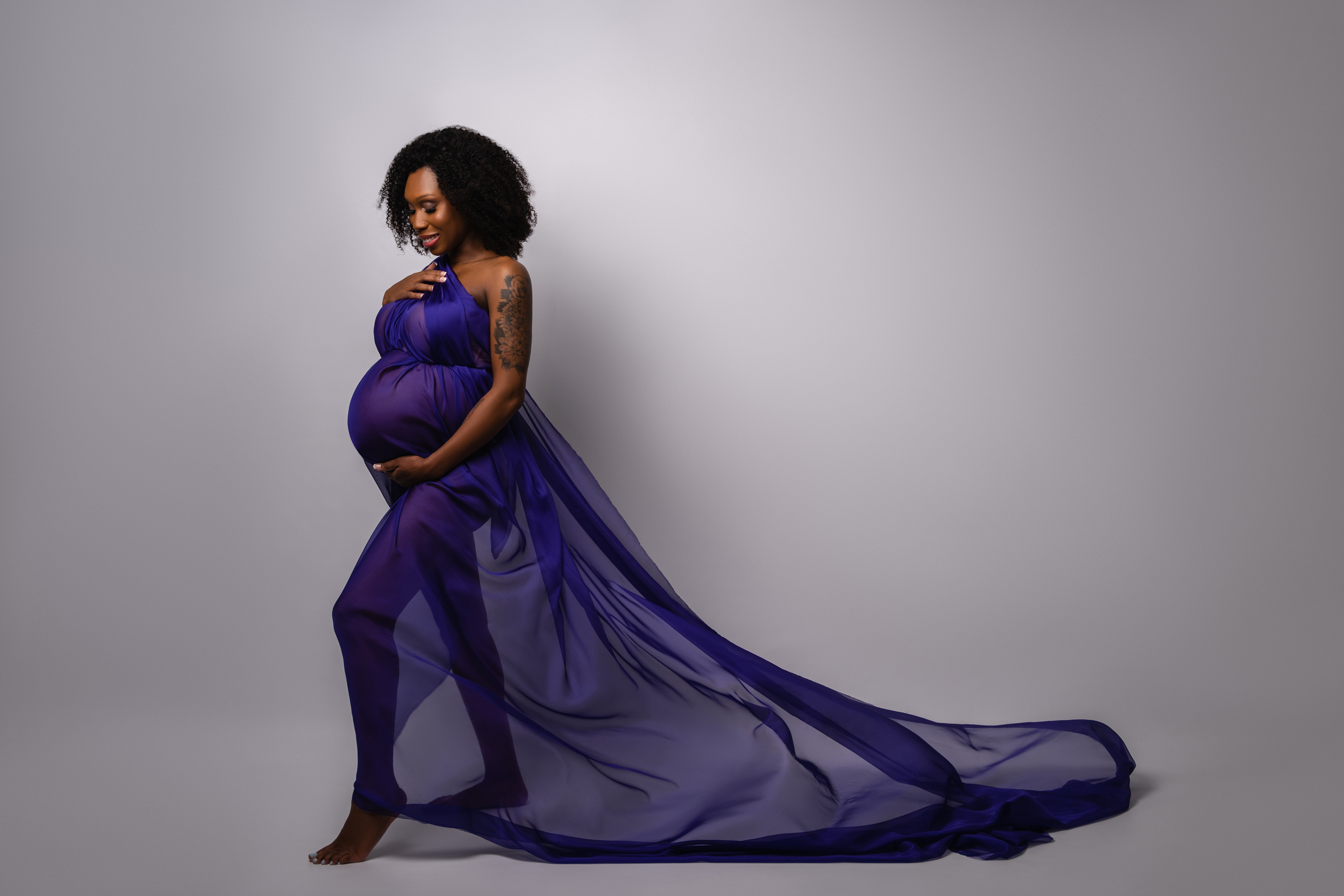 Wide maternity portrait in a flowing purple gown against a clean neutral studio background