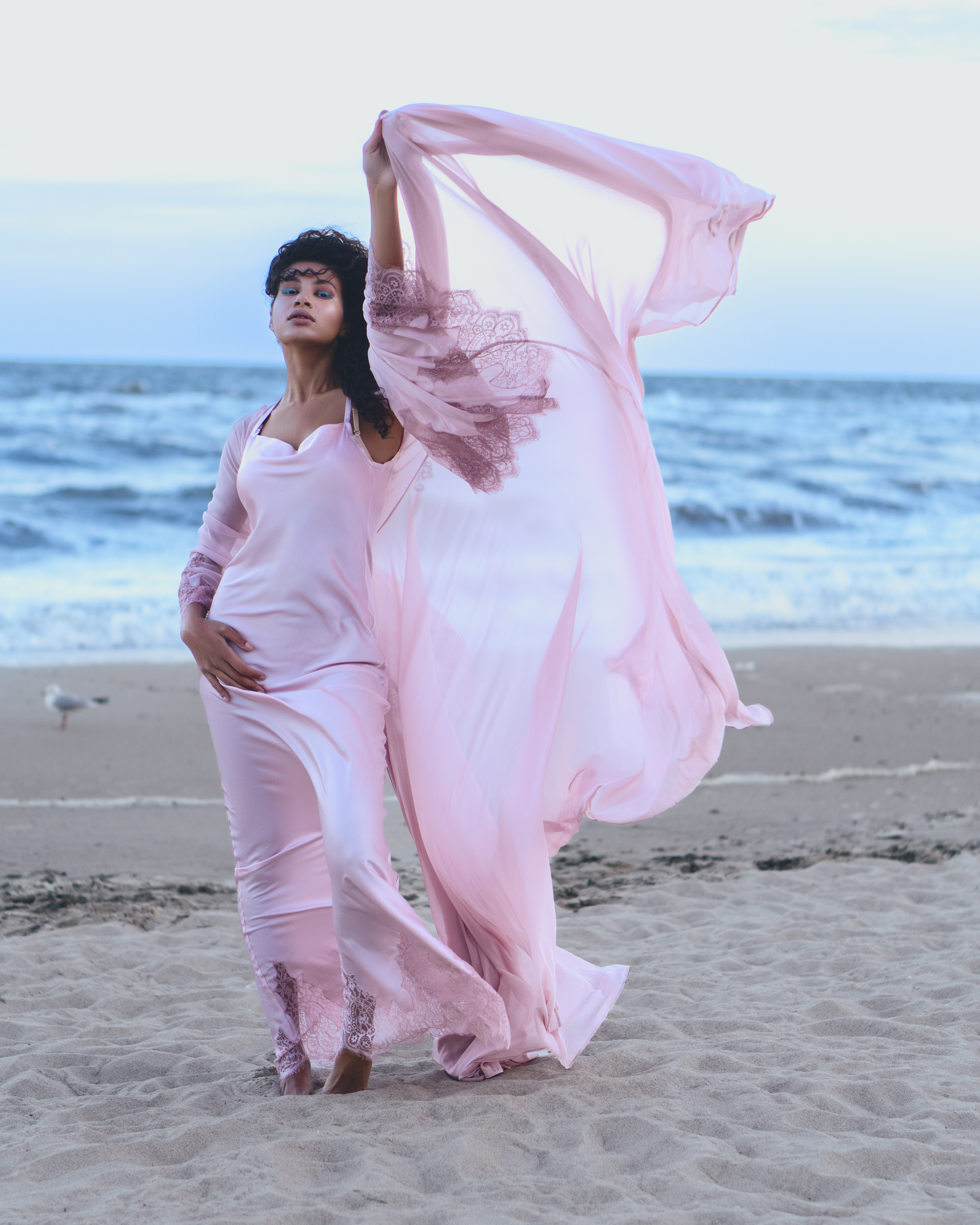 Coastal fashion portrait of a woman in a flowing blush-pink outfit posing on a beach with fabric lifted above her