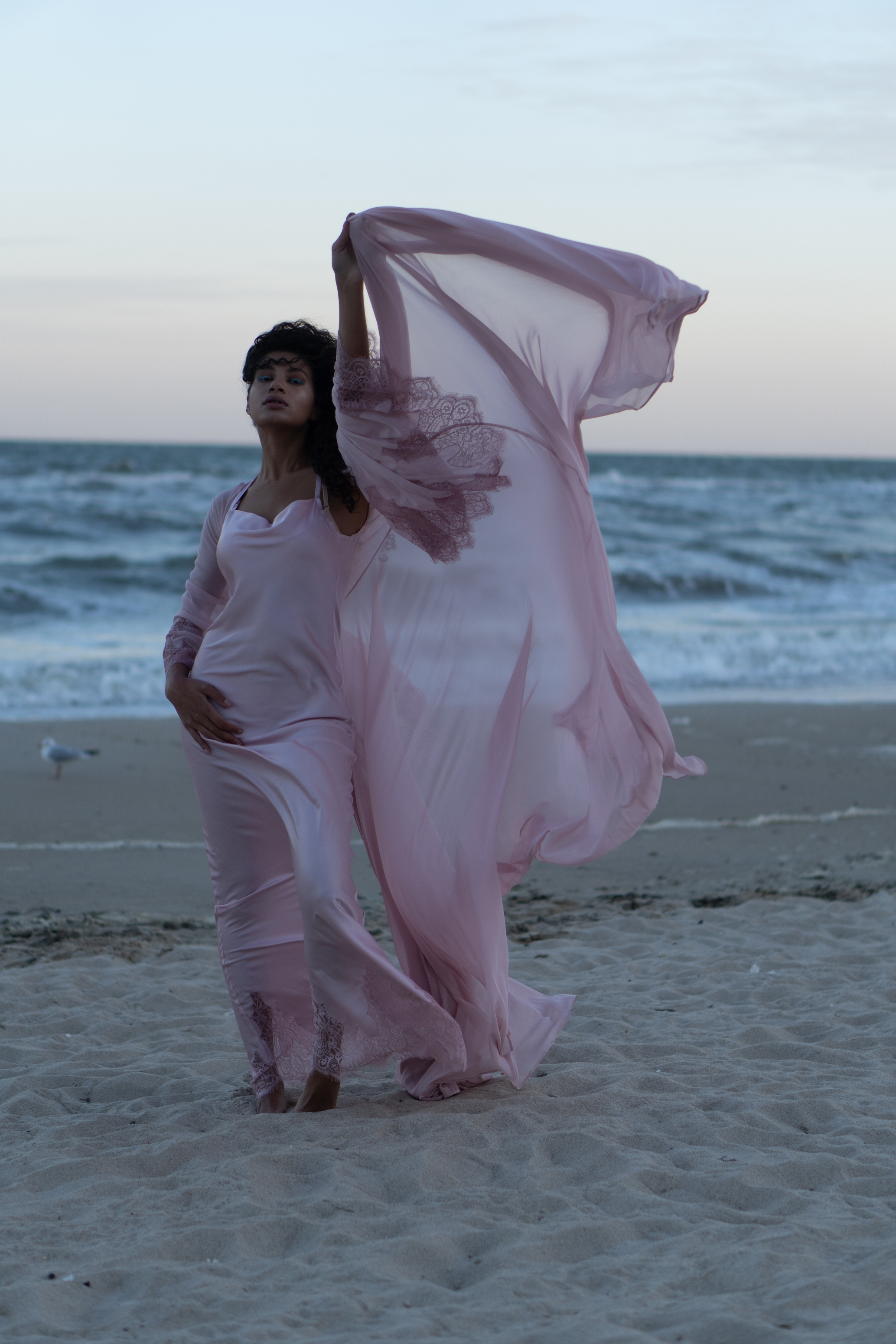 Raw full-length coastal fashion portrait of a woman in a pale pink flowing outfit standing on a beach