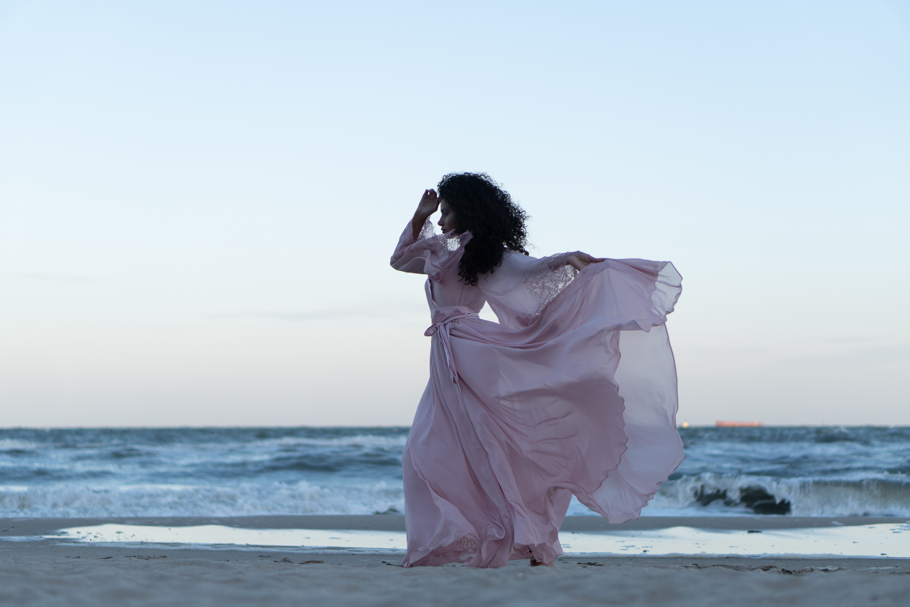 Raw wide fashion portrait of a woman in a flowing pink outfit walking along the beach